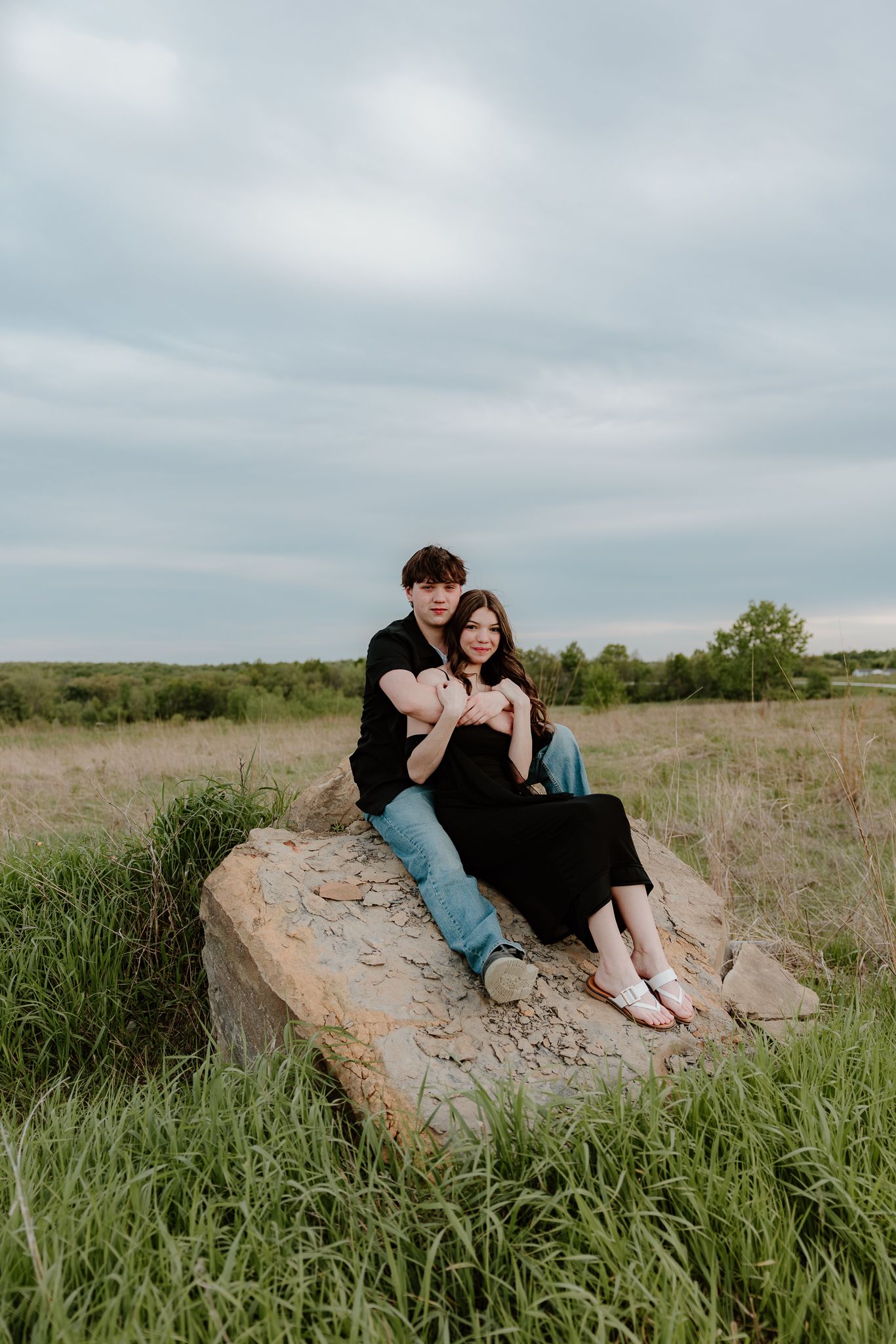 A couple sits closely on a large rock in a grassy field, the man in black and jeans embracing a woman in a black dress.