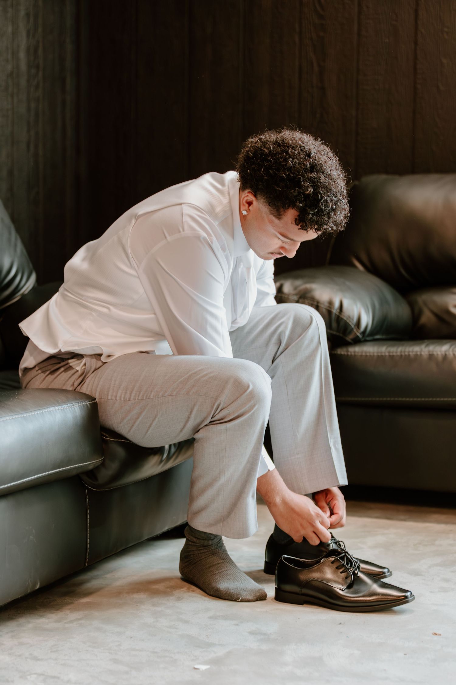 Groom in white suit sitting on leather sofa tying black dress shoes before wedding.