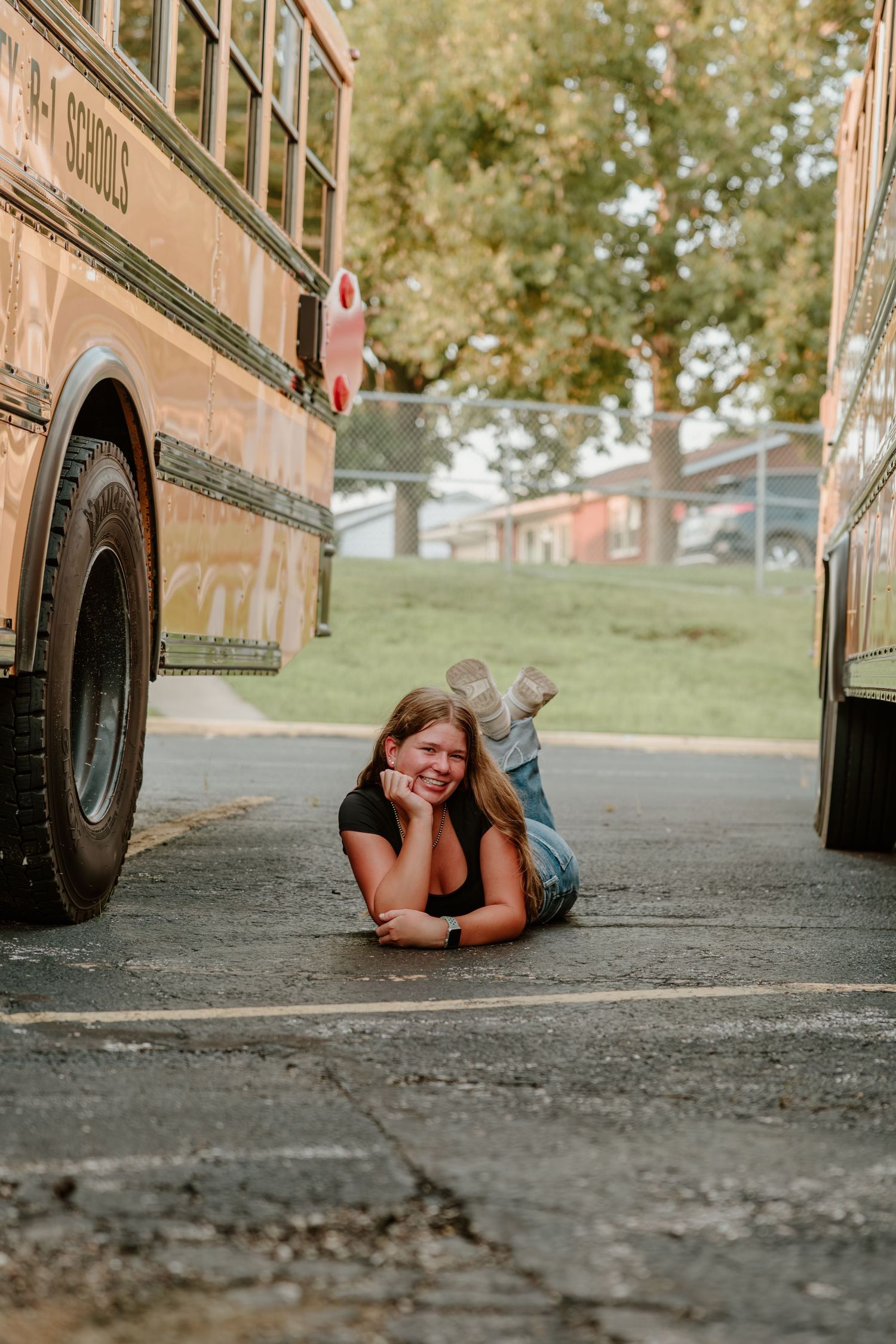 Someone laying playfully between two parked school buses on pavement with trees in the background.