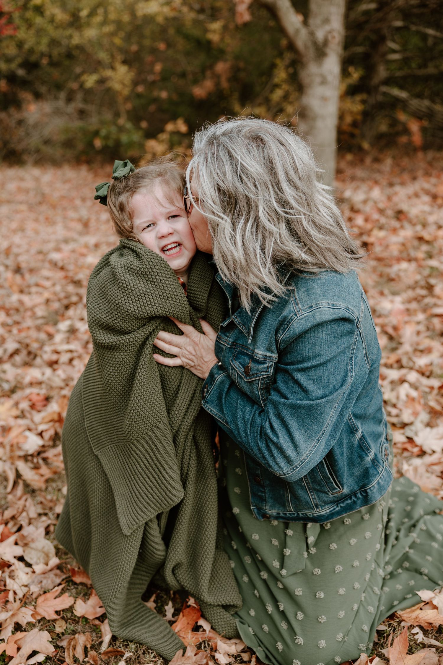 Two people embrace while sitting among fallen autumn leaves in a forest setting.