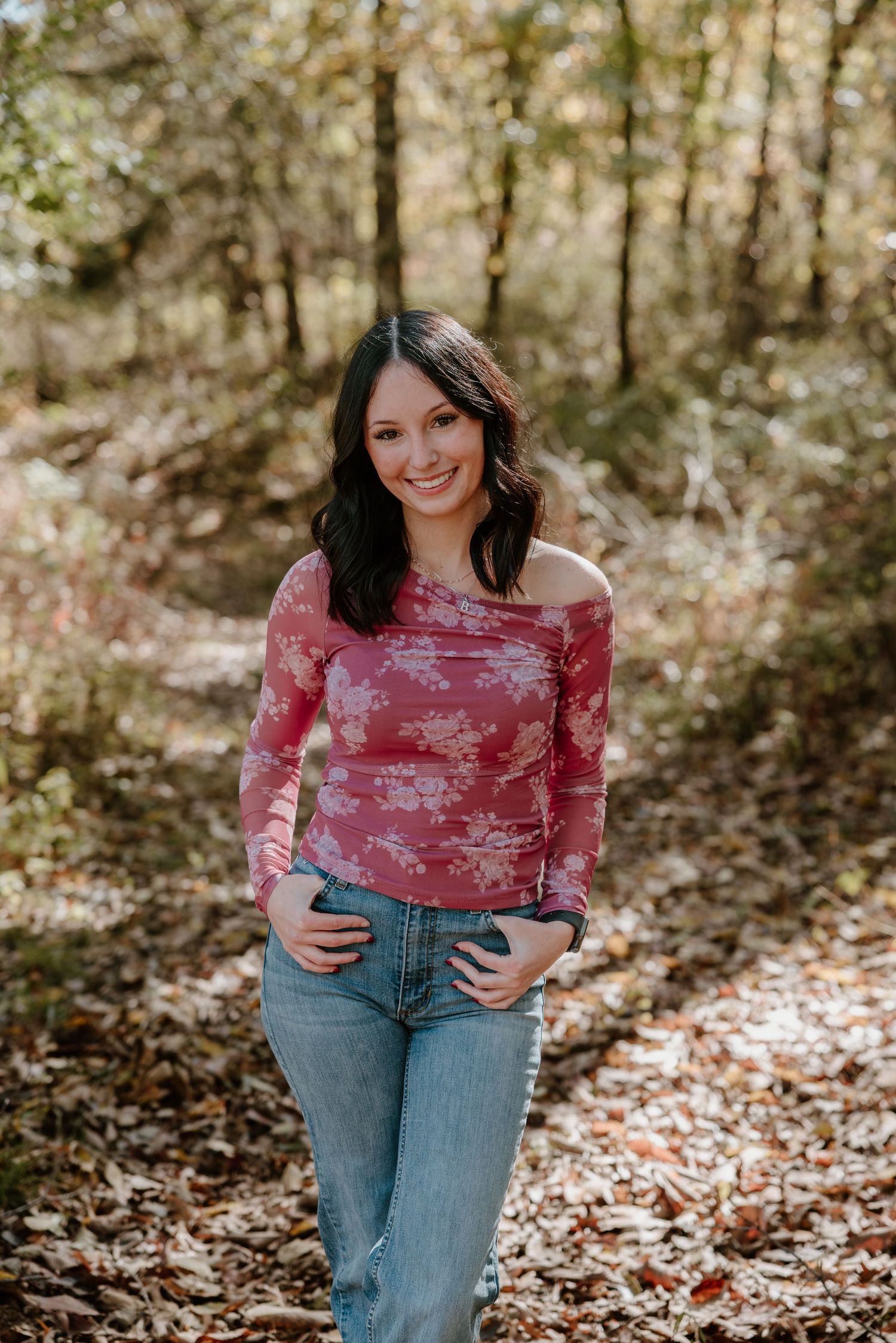 A woman in a pink floral top and jeans poses in a sunlit autumn forest setting.