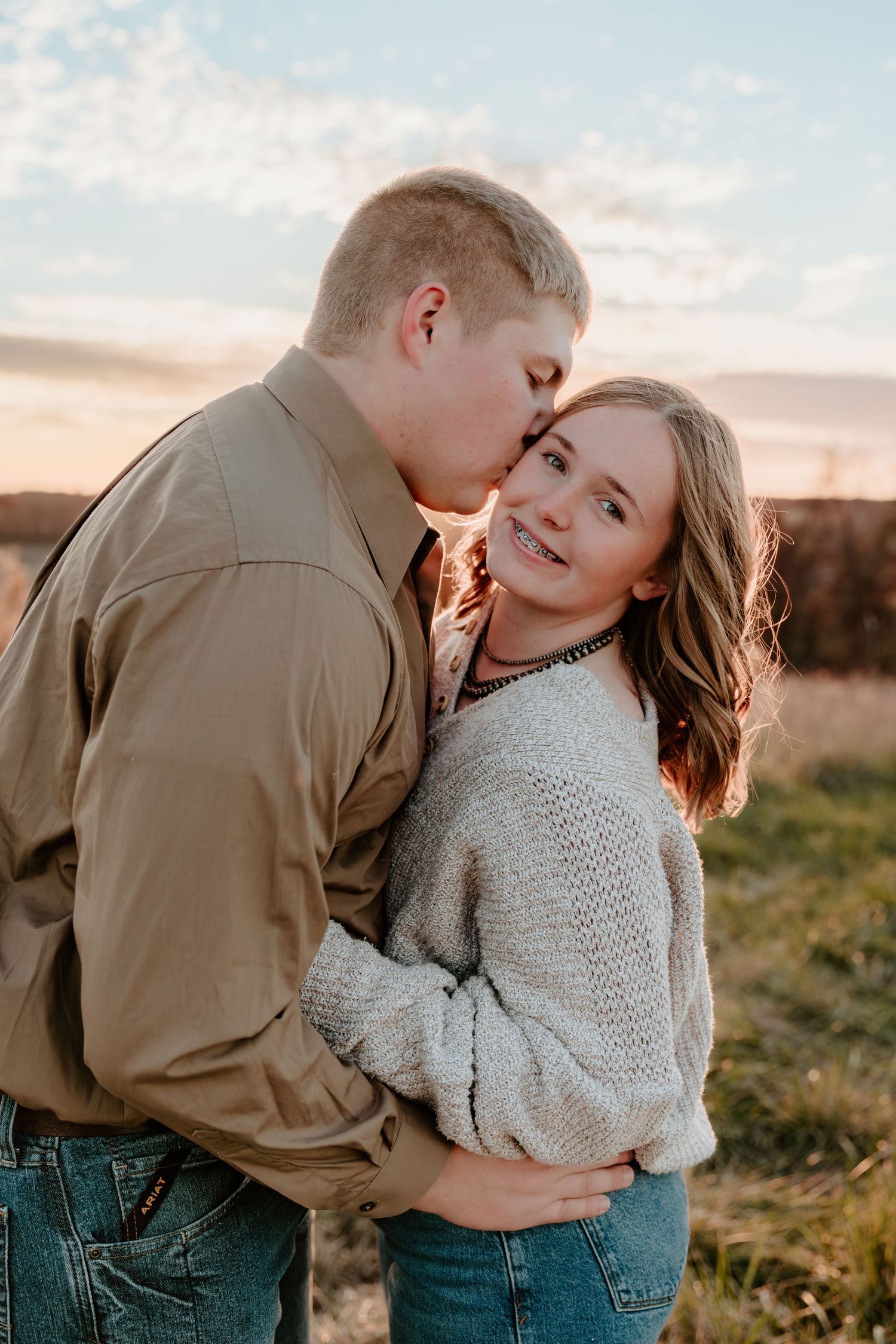 A romantic couple embraces and laughs together during a sunset photoshoot in a grassy outdoor field.