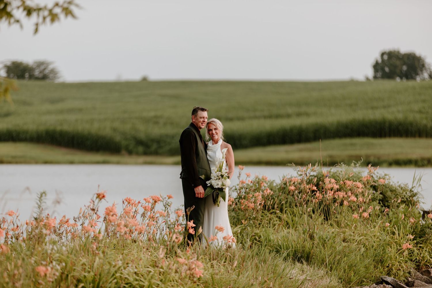 Bride and groom pose by a serene pond surrounded by wildflowers and lush green fields on their wedding day.