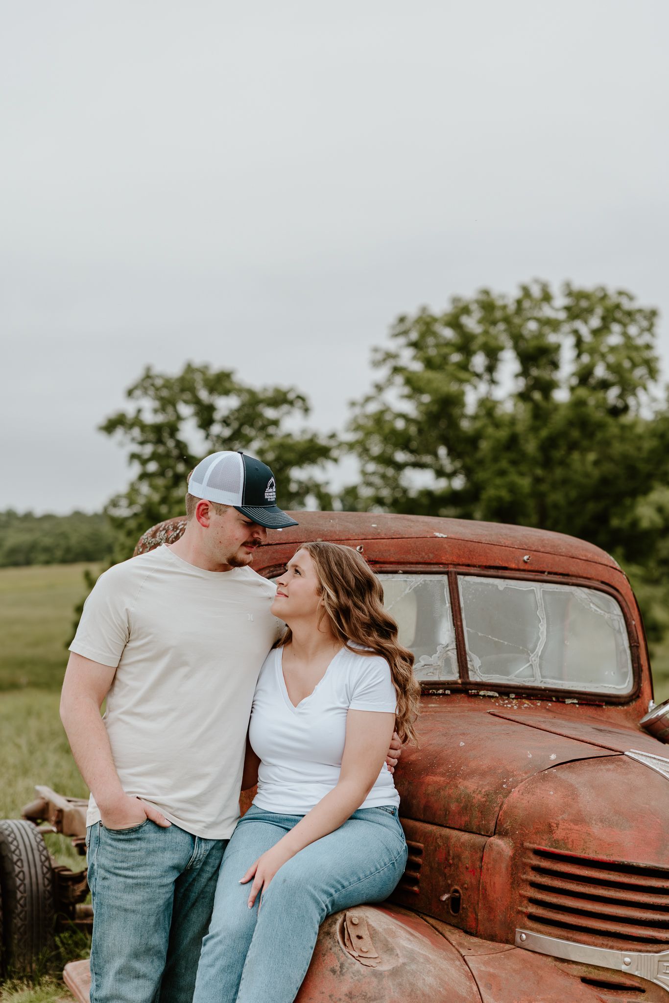 Couple leaning against a rustic red vintage truck in a green field, wearing white tees and jeans.