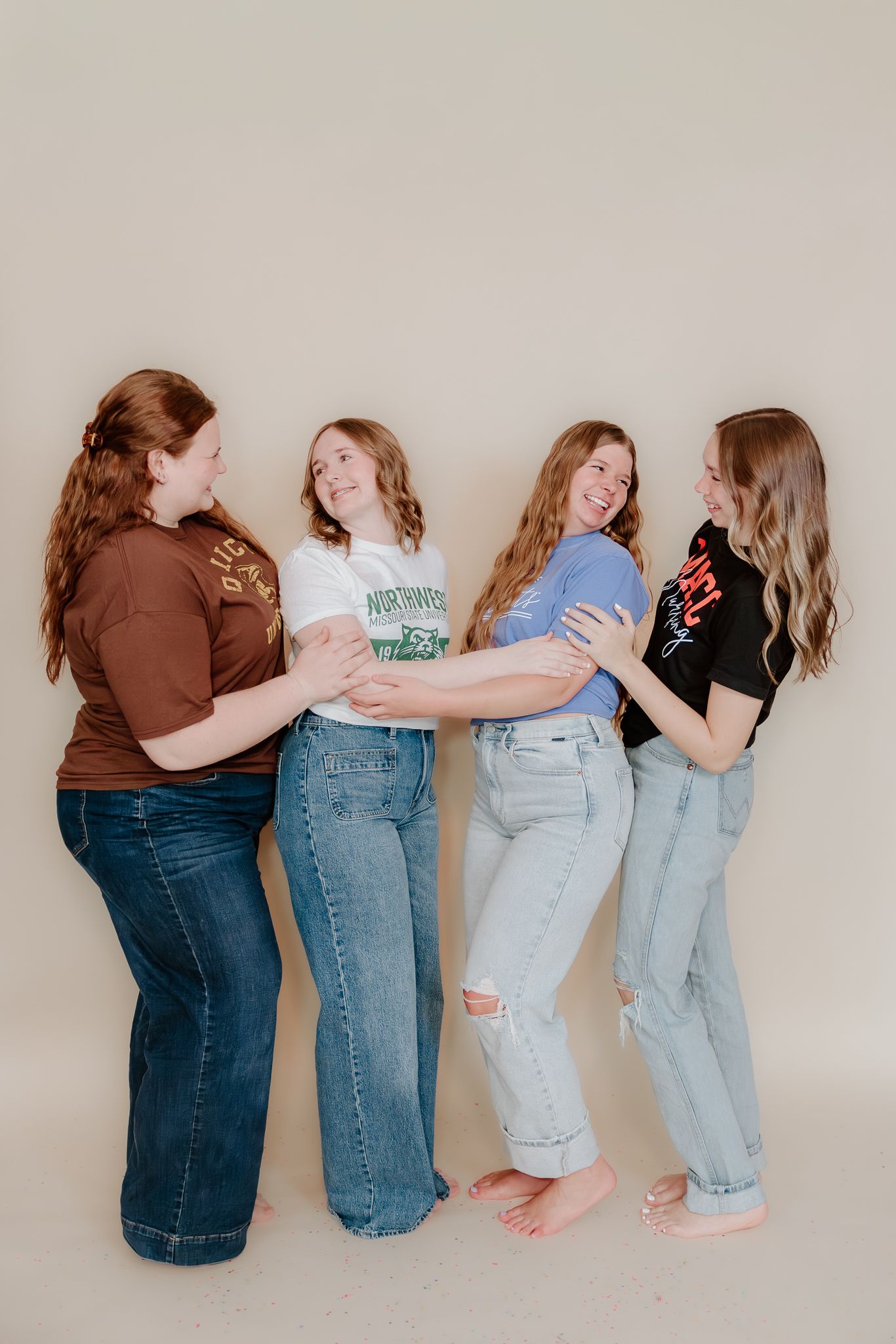 Four young women laughing and posing together in casual jeans and graphic tees against a neutral beige background.