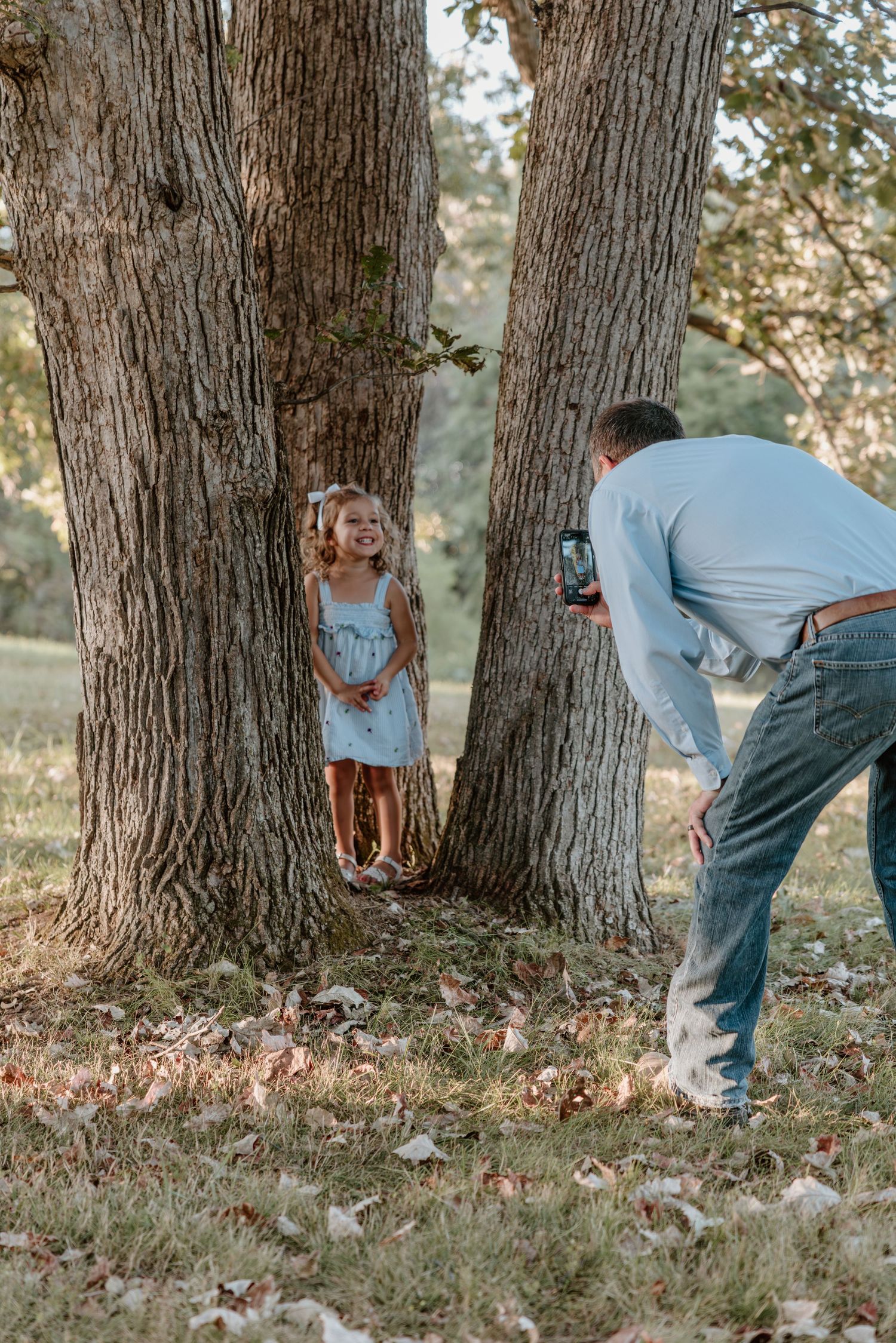 A playful sequence of peek-a-boo moments around a large tree trunk in a park setting with autumn leaves on the ground.