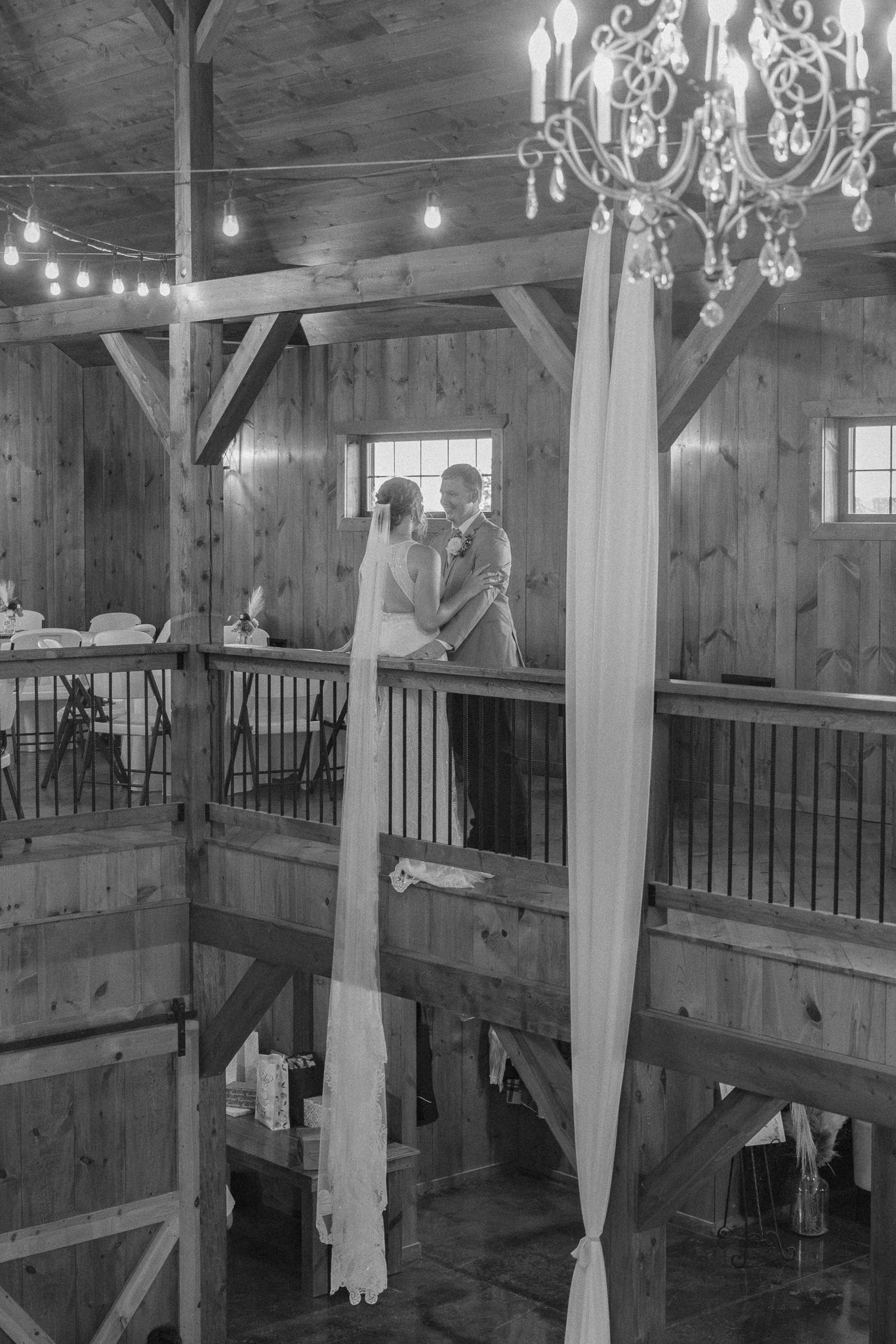 Black and white photo of a bride and groom embracing on a rustic barn loft with chandeliers and string lights.