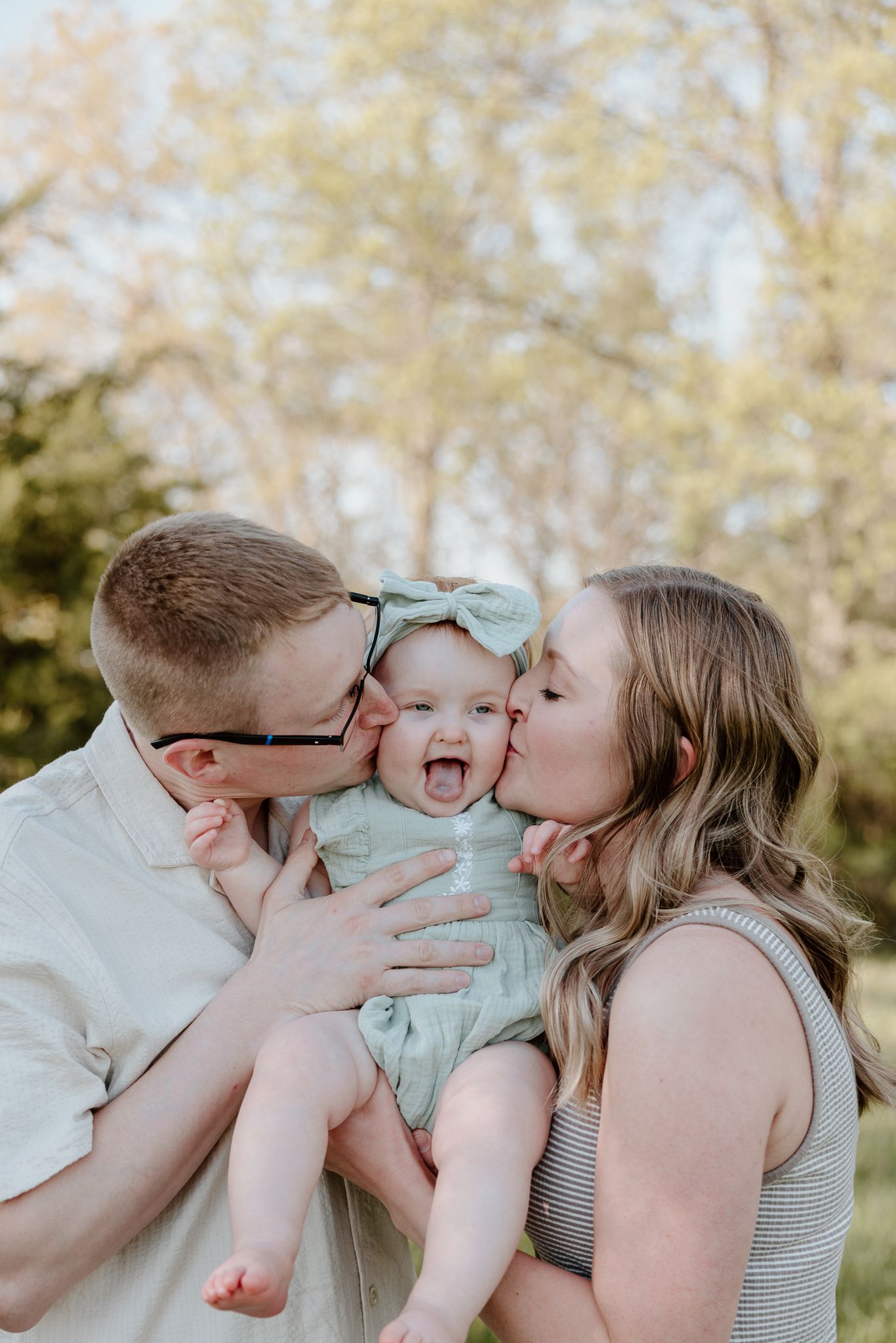 Happy baby laughing as mom and dad kiss her cheeks in a sunny outdoor family portrait.