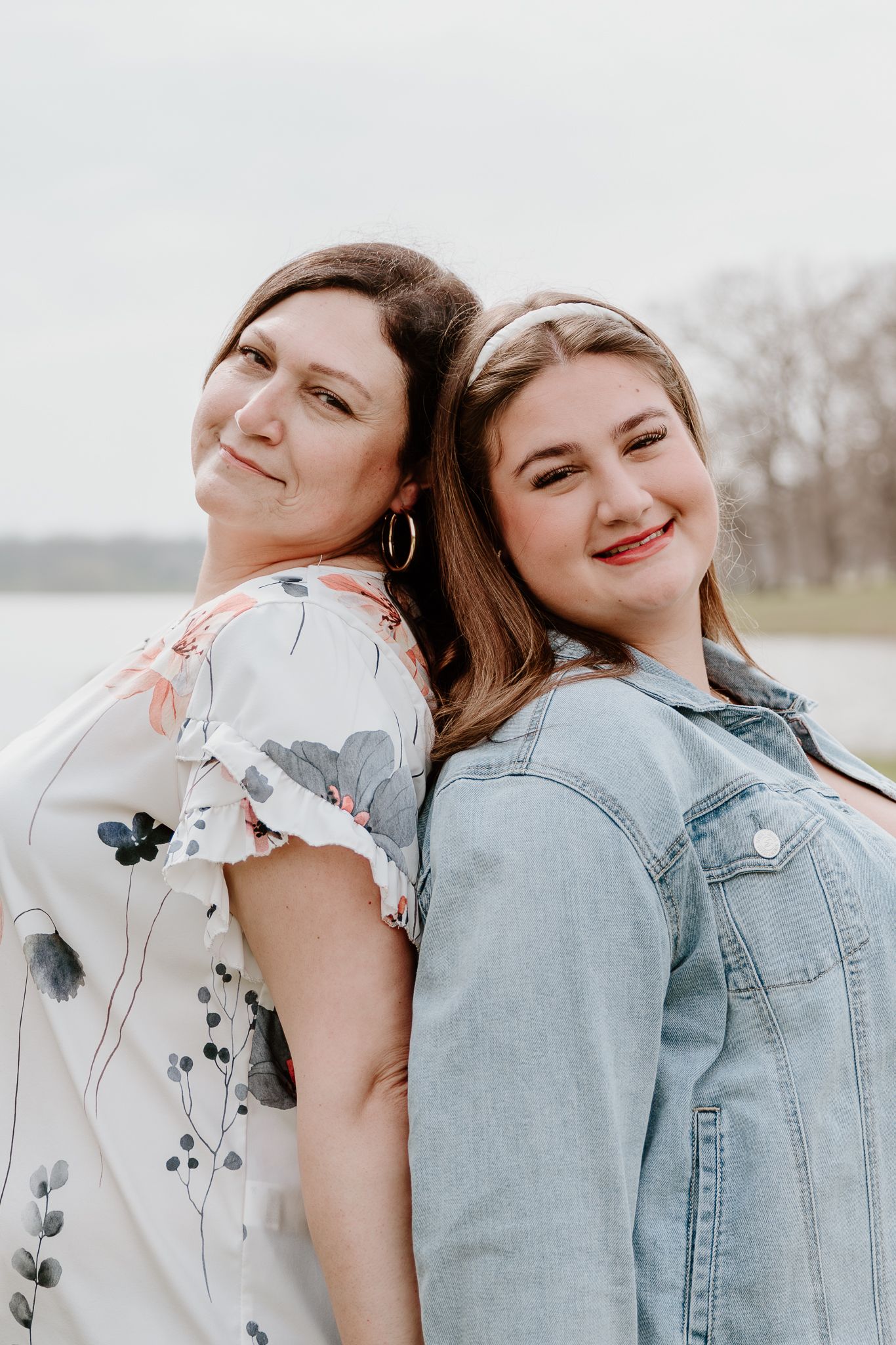 Two women smile back-to-back outdoors near a lake, one in a floral dress and one in a denim jacket.