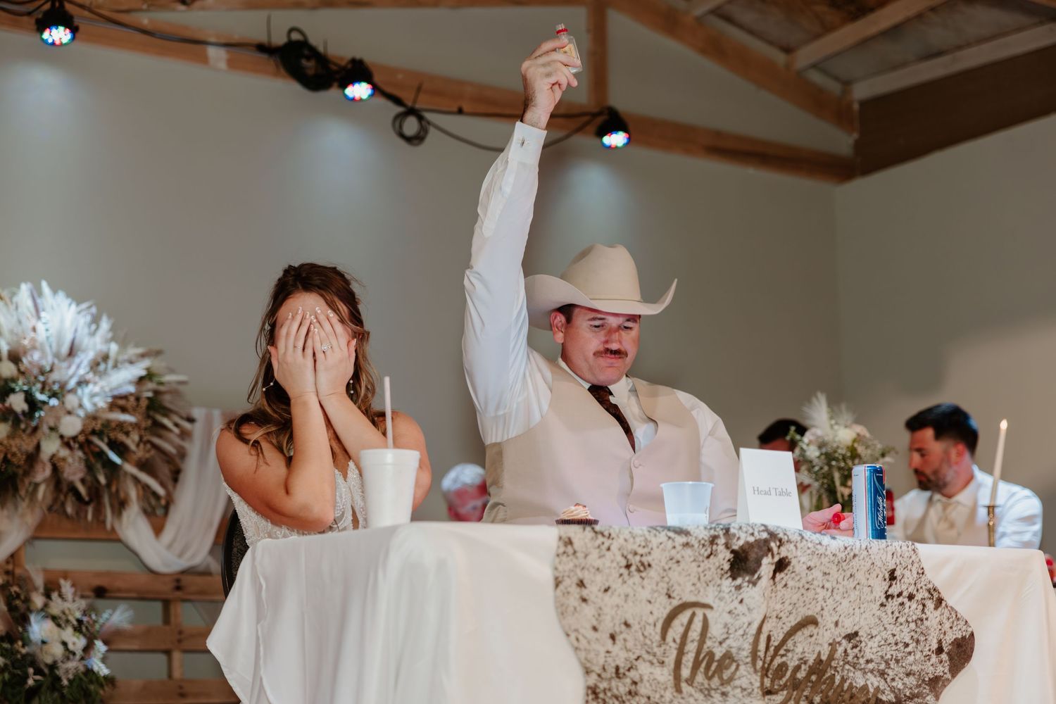 Bride covers face while groom in cowboy hat raises hand in celebration at rustic wedding reception table.