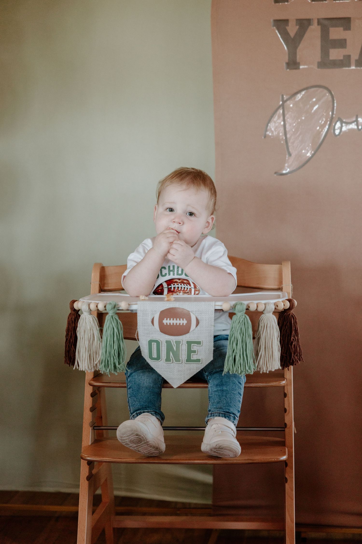 Baby sits in wooden high chair wearing white shirt and jeans with football and one year birthday decorations.
