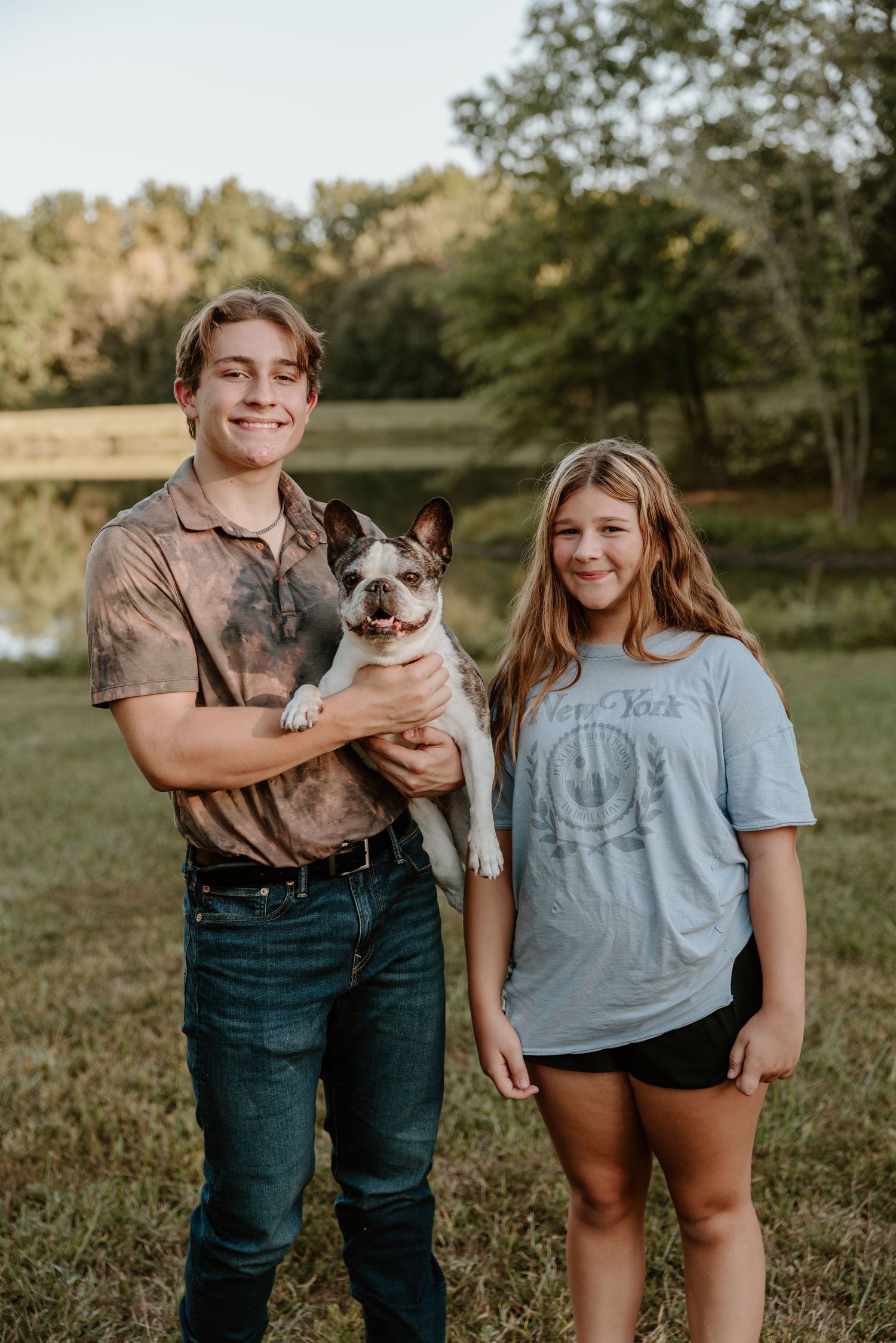 A couple stands outdoors in a grassy field at sunset while holding their Boston Terrier dog.