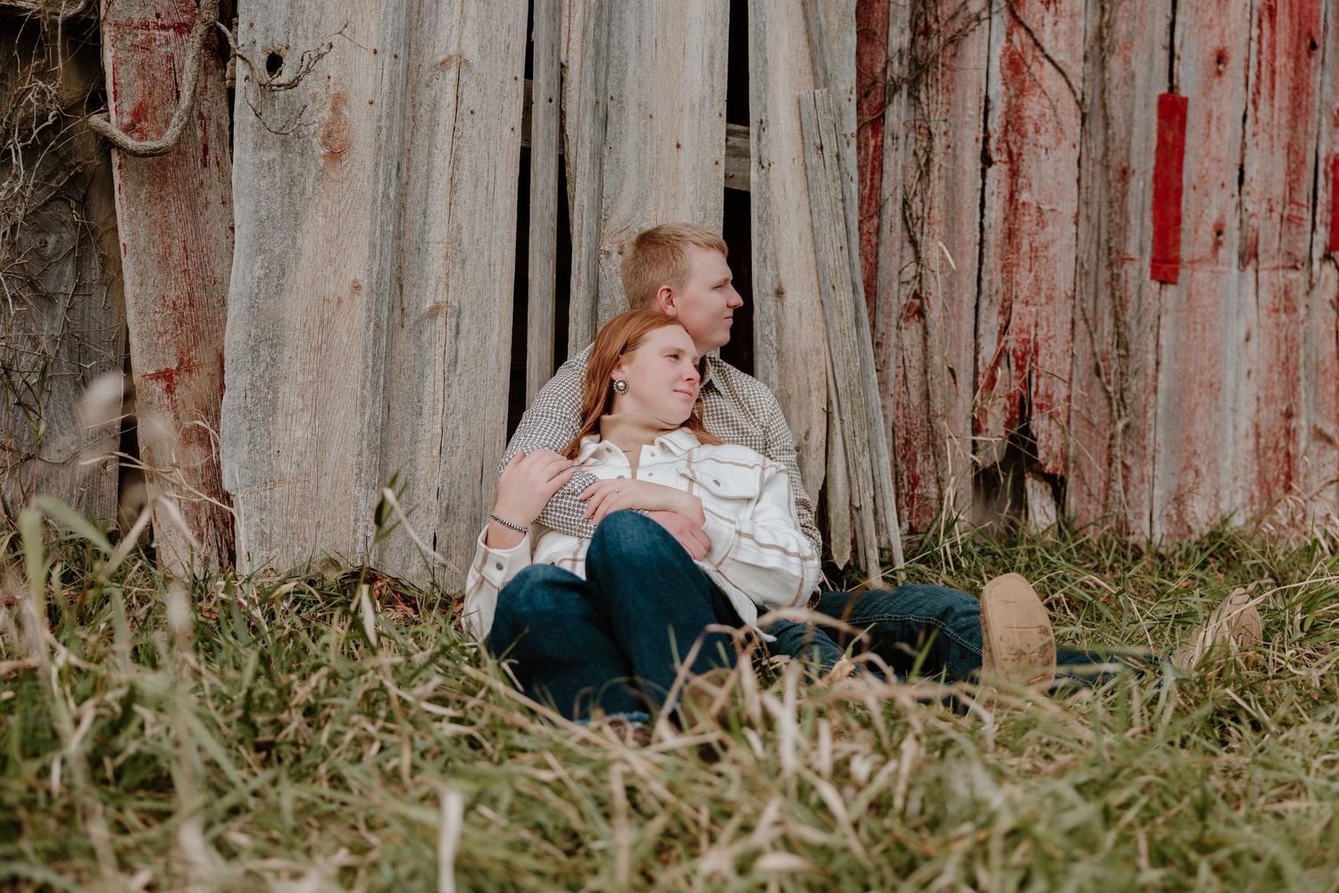 A romantic couple shares an intimate moment while sitting in tall grass against a rustic wooden fence.