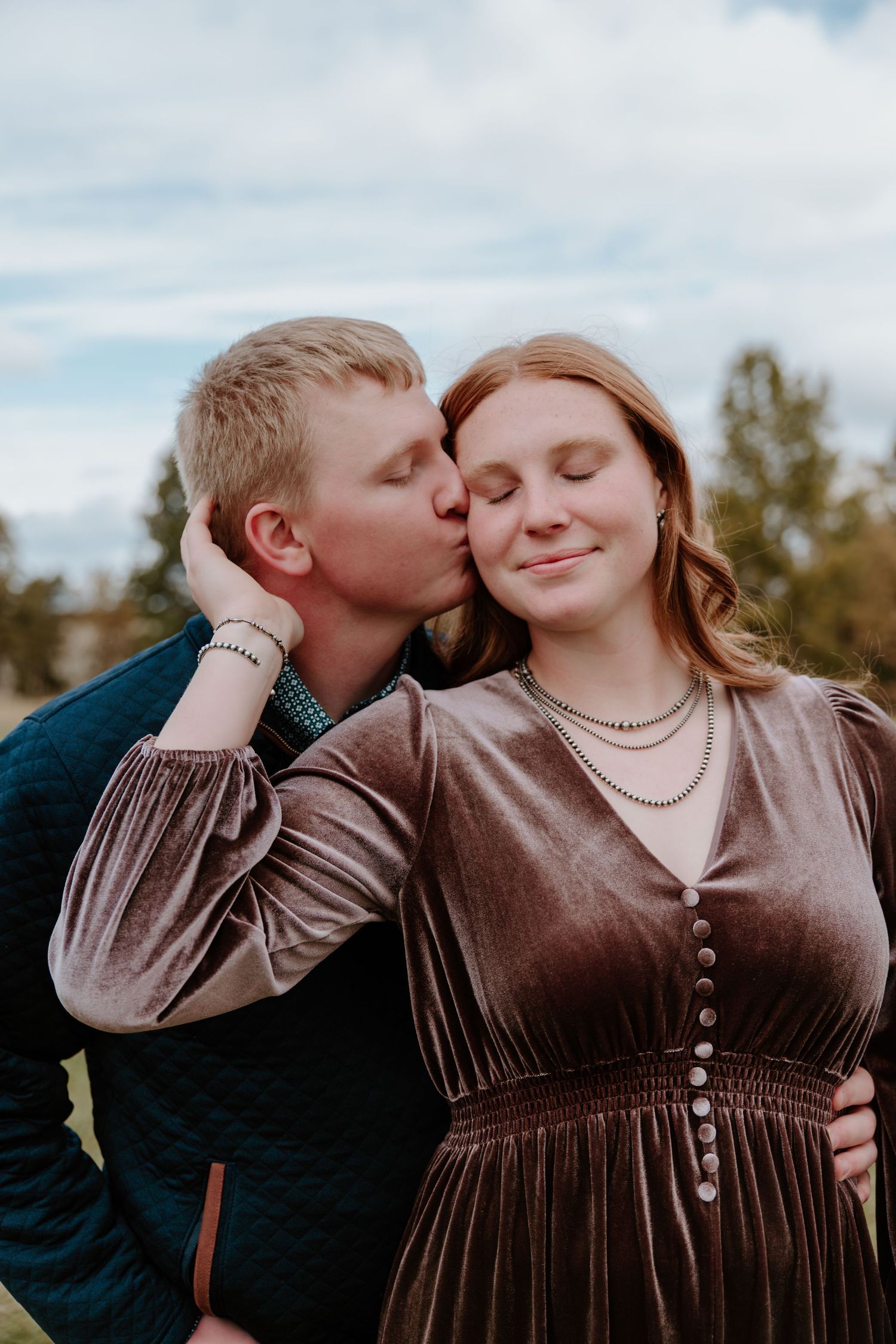 A romantic couple in brown dress and dark shirt share tender moments outdoors under a cloudy sky.
