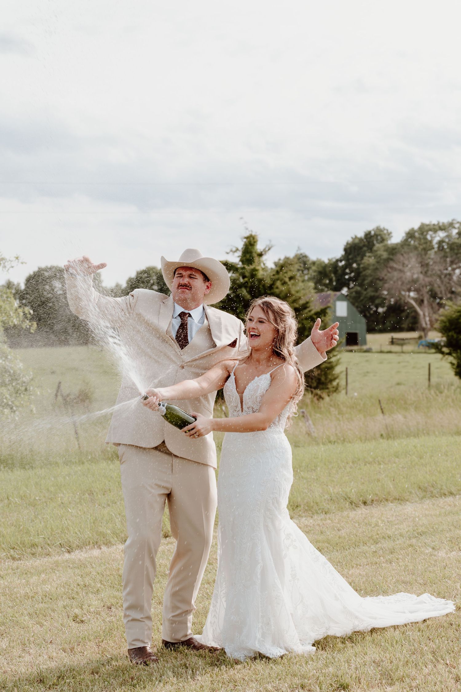 Joyful bride and groom in cowboy hat popping champagne bottle outdoors on green lawn at rustic country wedding.