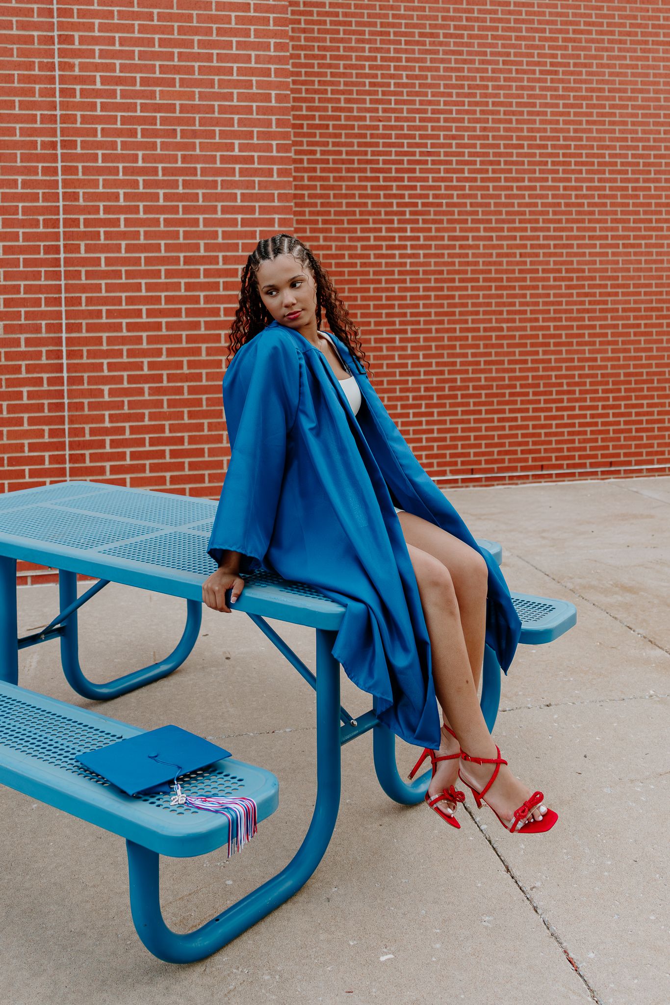 A young woman in a blue graduation gown and red heels sits on a blue picnic table against a brick wall.