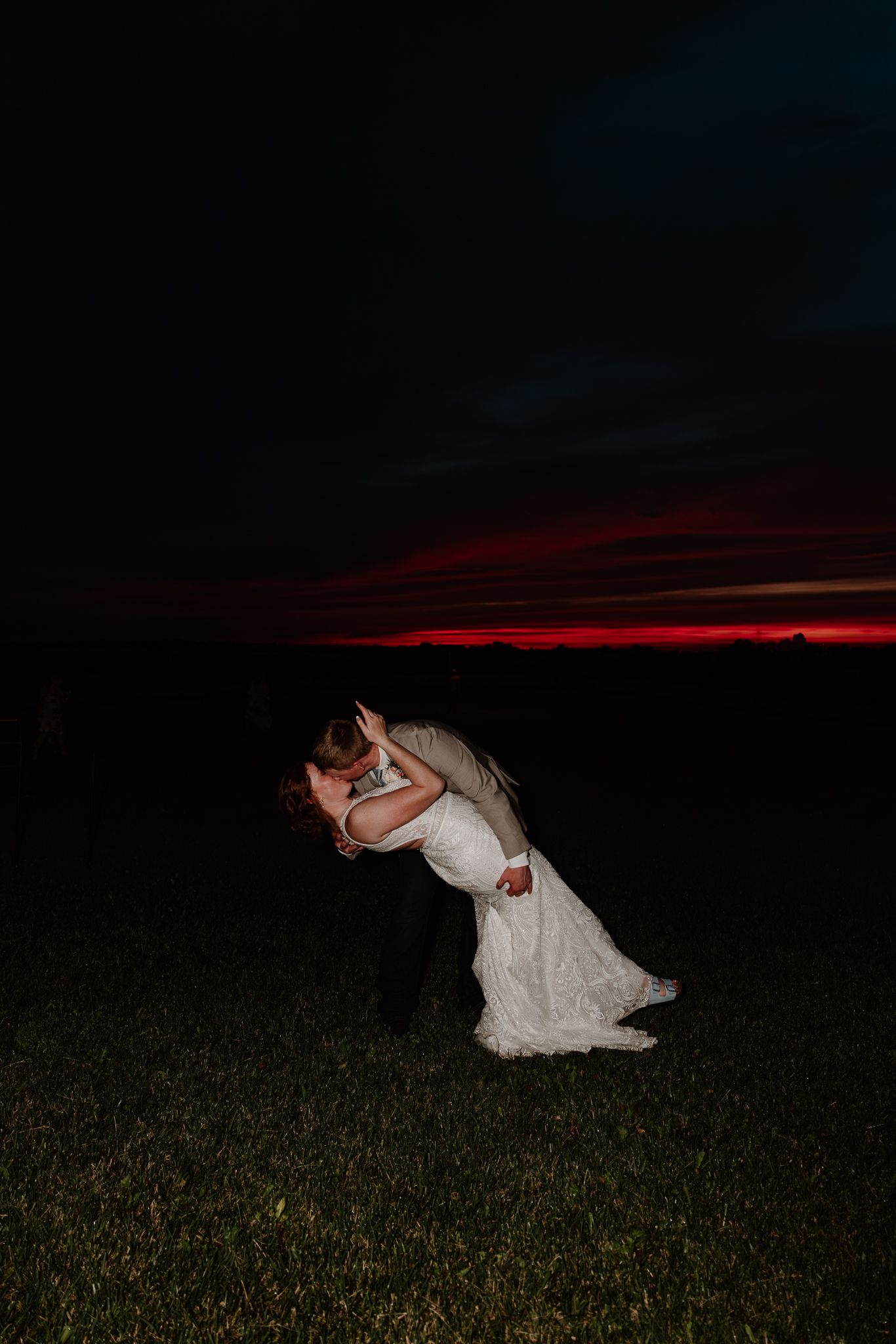 Bride and groom share a romantic dip kiss at sunset on a grassy field with a vivid red horizon.