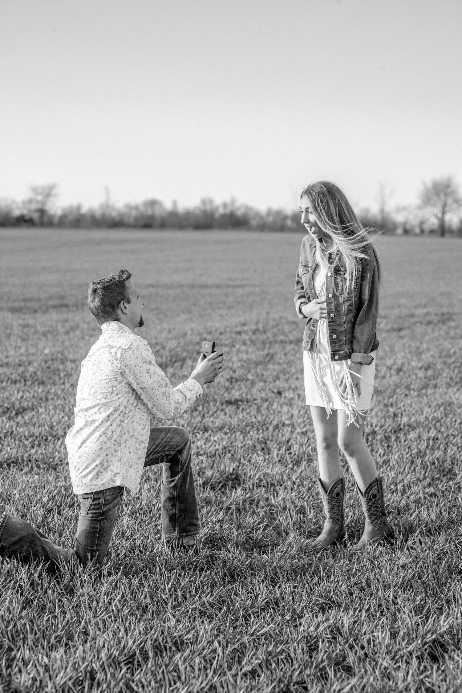 Marriage proposal moment captured in black and white on grassy field.
