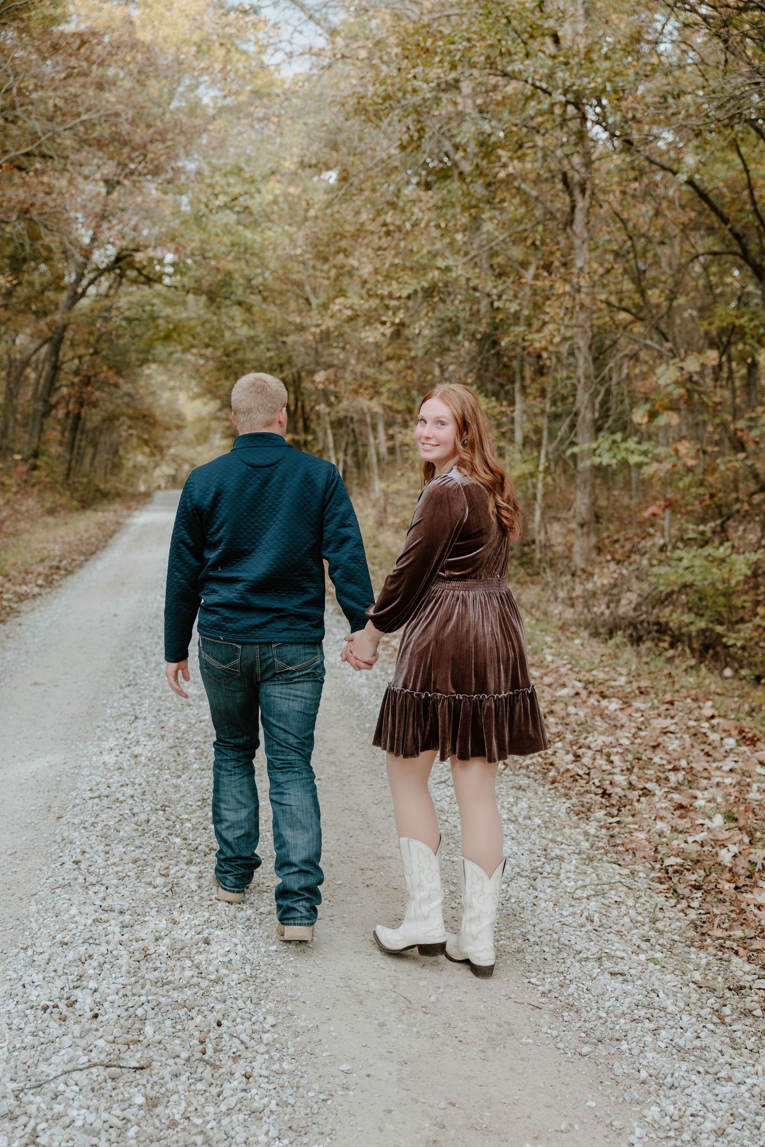 A couple walks hand in hand down a gravel path surrounded by autumn trees in a romantic outdoor setting.