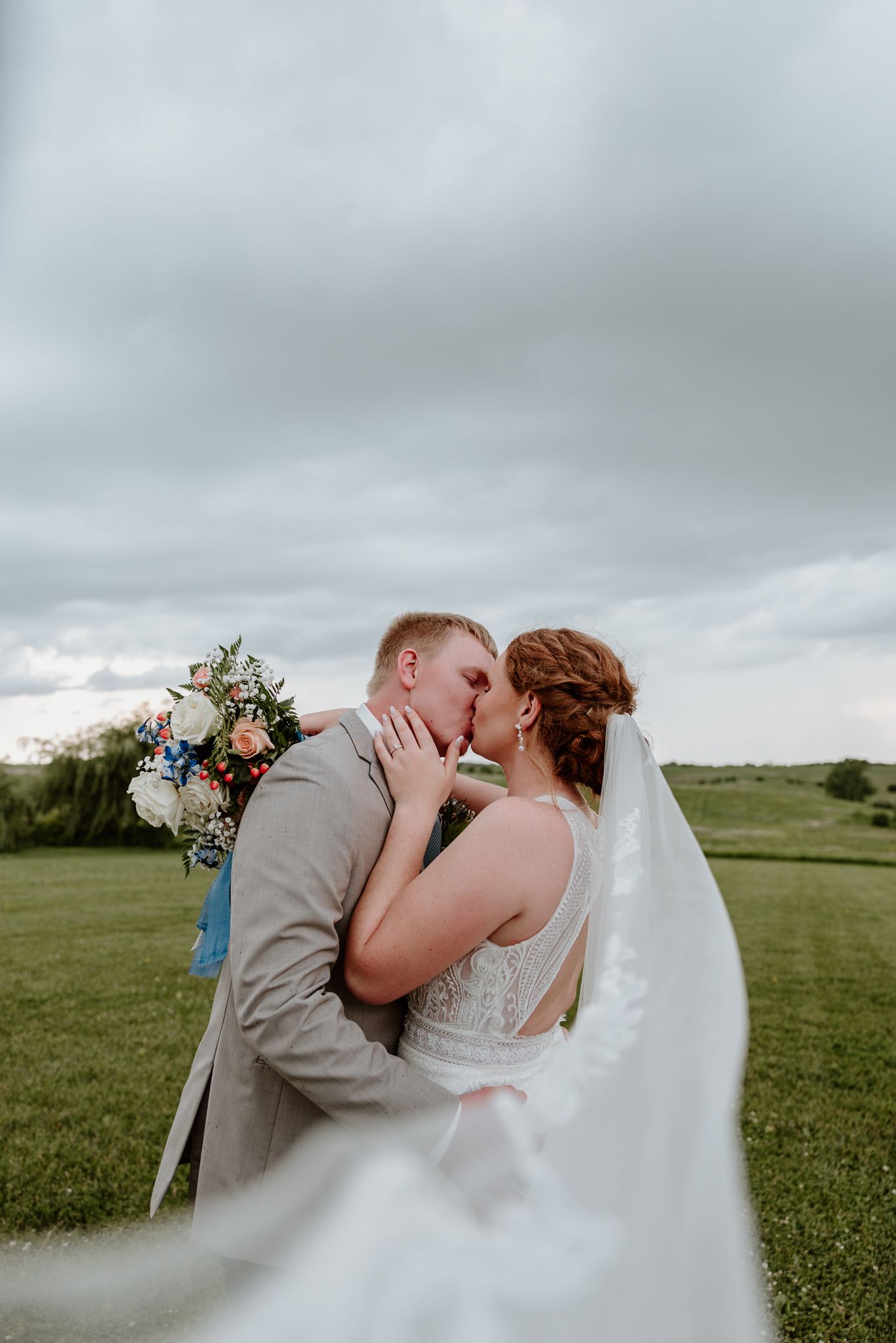 Bride and groom sharing a romantic kiss outdoors with veil flowing in the wind on green field.