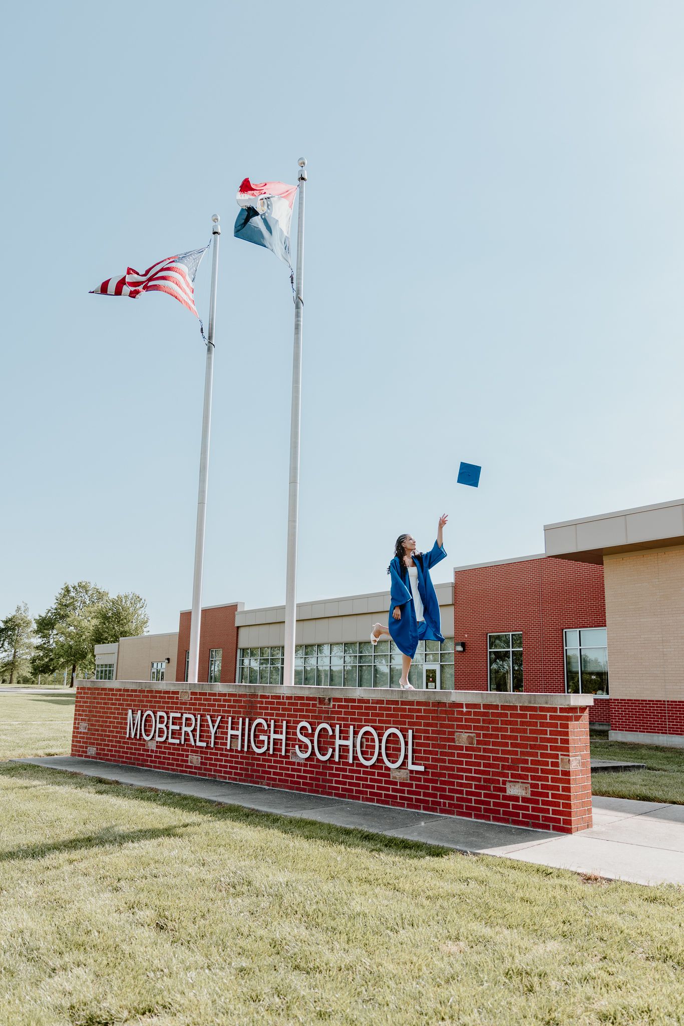 Graduate in blue cap and gown tosses cap in front of Moberly High School sign with flags waving.