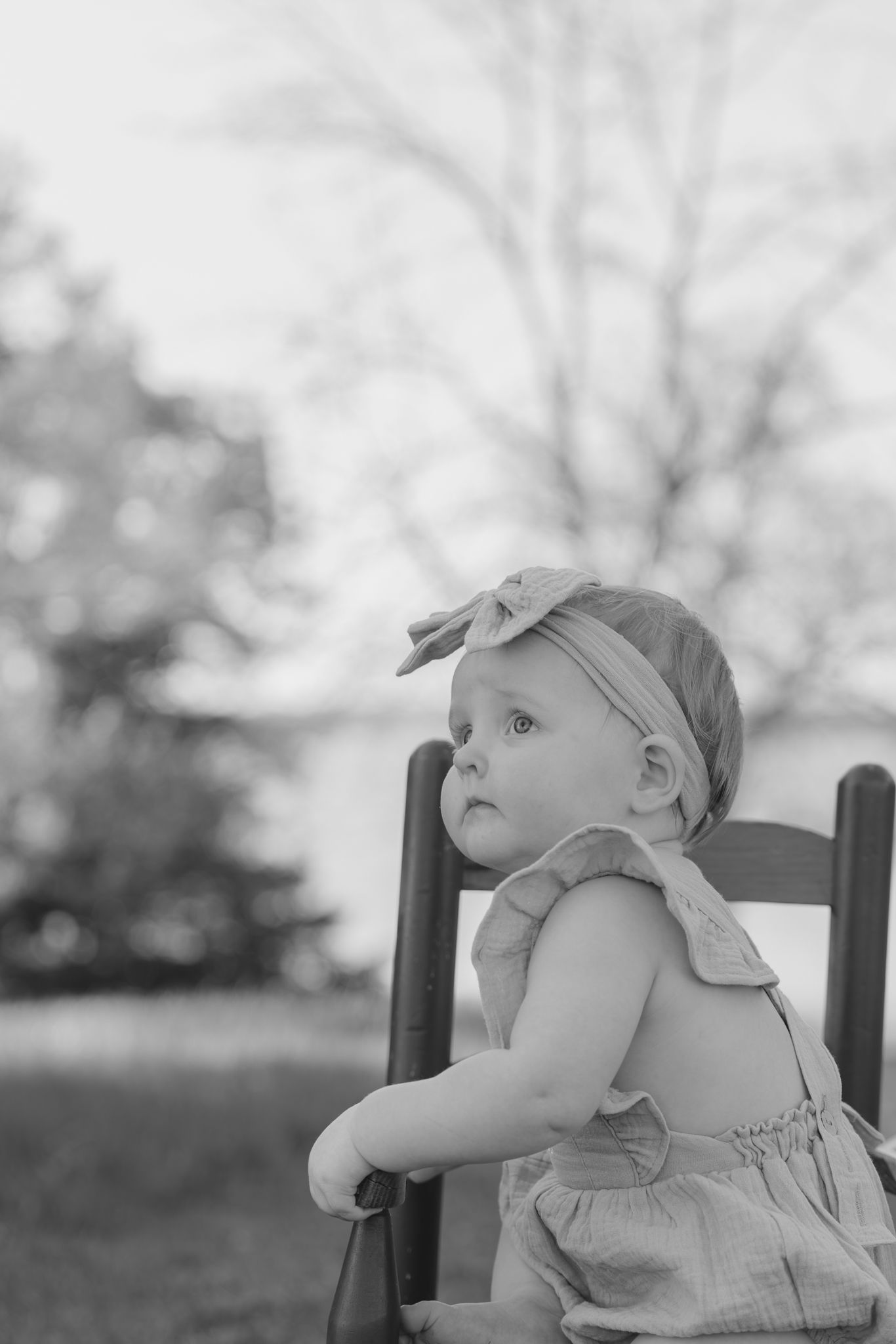 Black and white photo of a cute baby girl in a romper and bow headband sitting on a wooden chair outdoors.