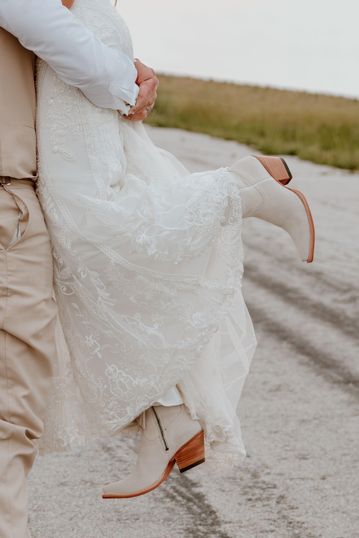 A bride in a lace wedding dress and white cowboy boots is lifted by her groom on a scenic outdoor road.