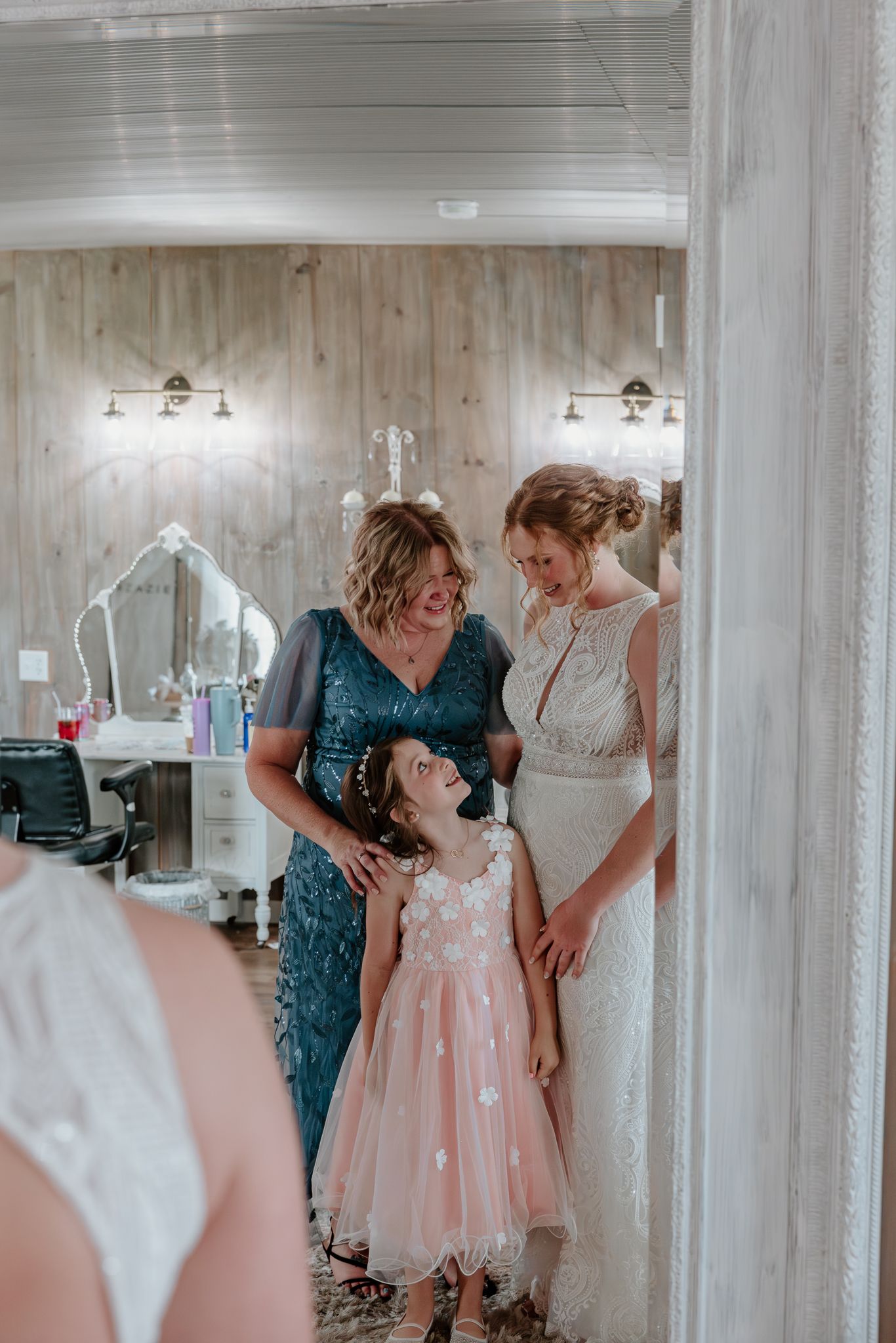 Bride in white lace gown shares a joyful moment with woman in teal dress and flower girl in pink tulle dress.
