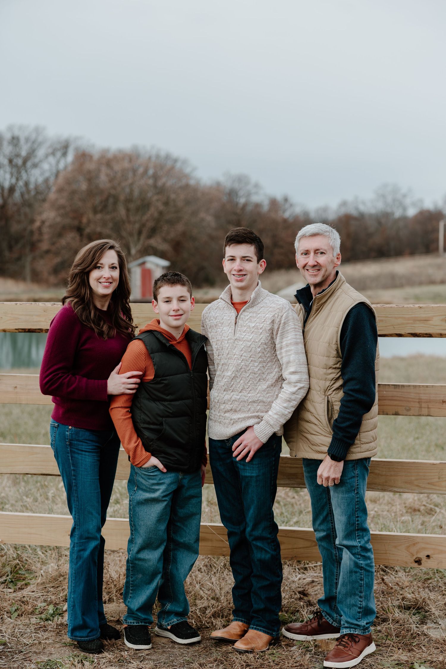 Family of four poses together by wooden fence at pond wearing casual sweaters and jeans on overcast autumn day.
