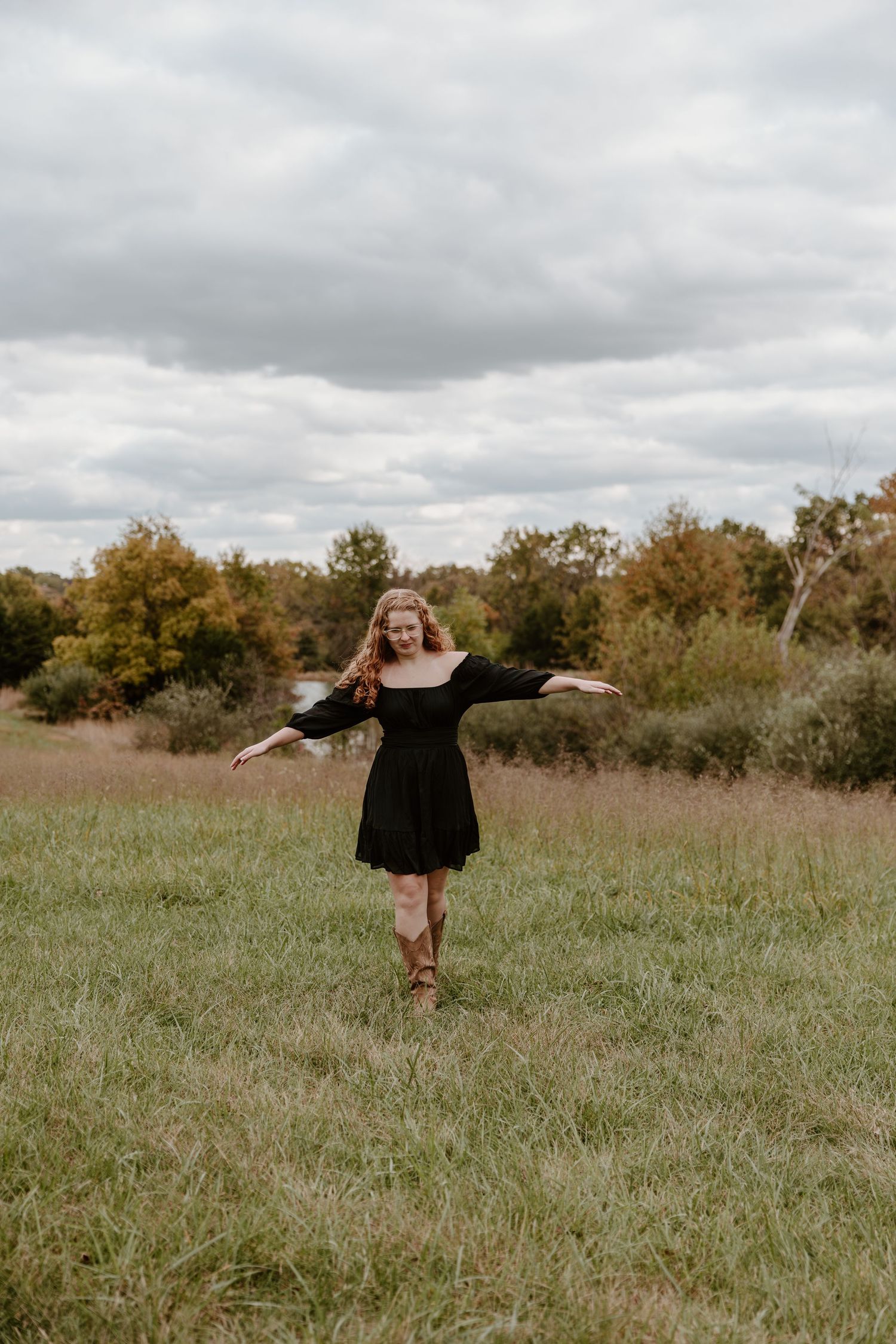 A person in a black dress twirls joyfully in a rustic autumn field under cloudy skies.