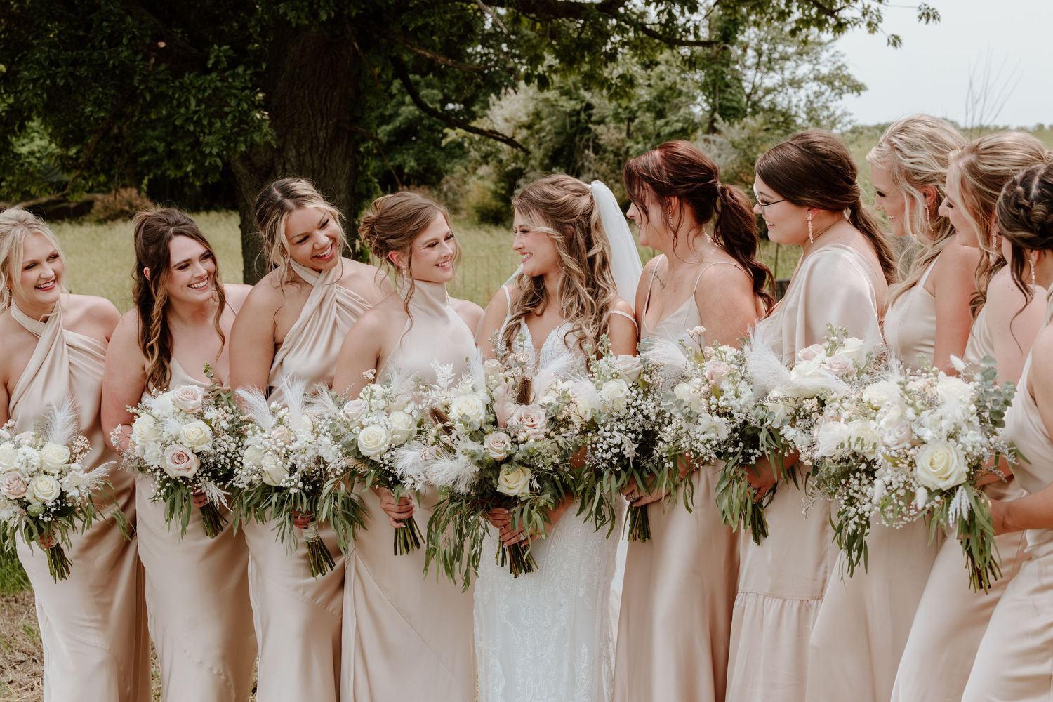 Bride and bridesmaids in blush dresses laughing together holding white floral bouquets outdoors.