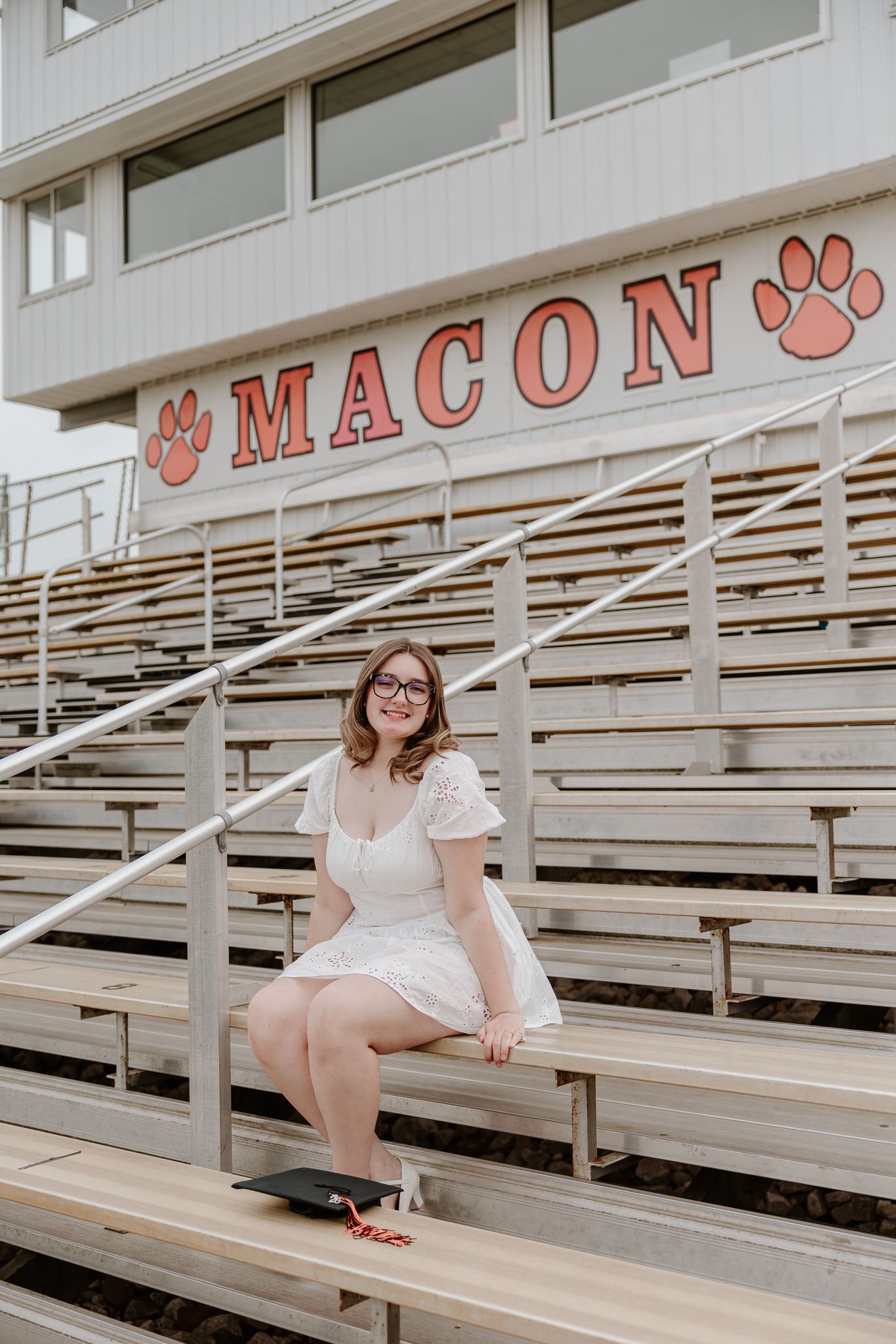 Young woman in white dress sitting on bleachers at Macon school stadium, smiling with graduation cap nearby.