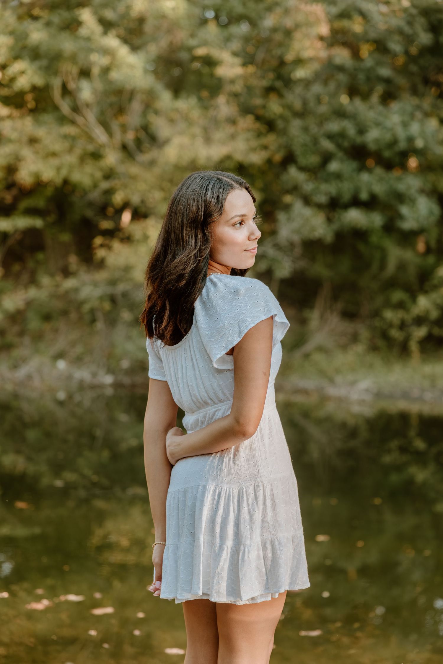 A person in a white summer dress poses gracefully near a pond in a natural outdoor setting.