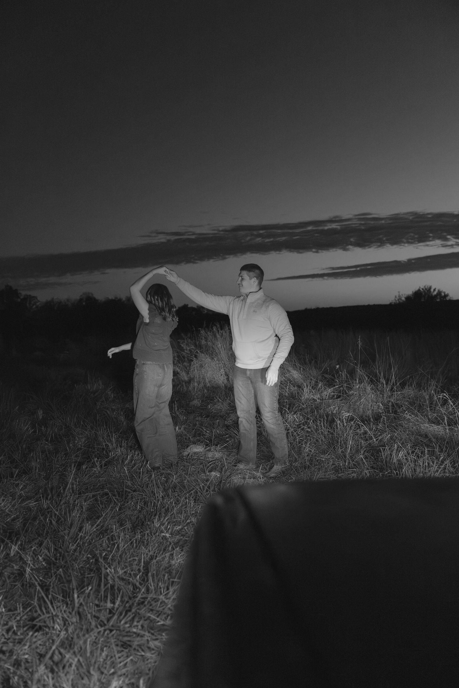 A silhouetted couple dancing playfully in a field against a dramatic sunset sky.