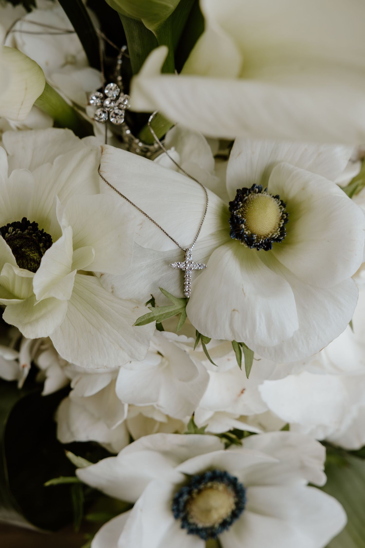 Silver cross necklace and earrings resting on white anemone flowers in bridal bouquet.