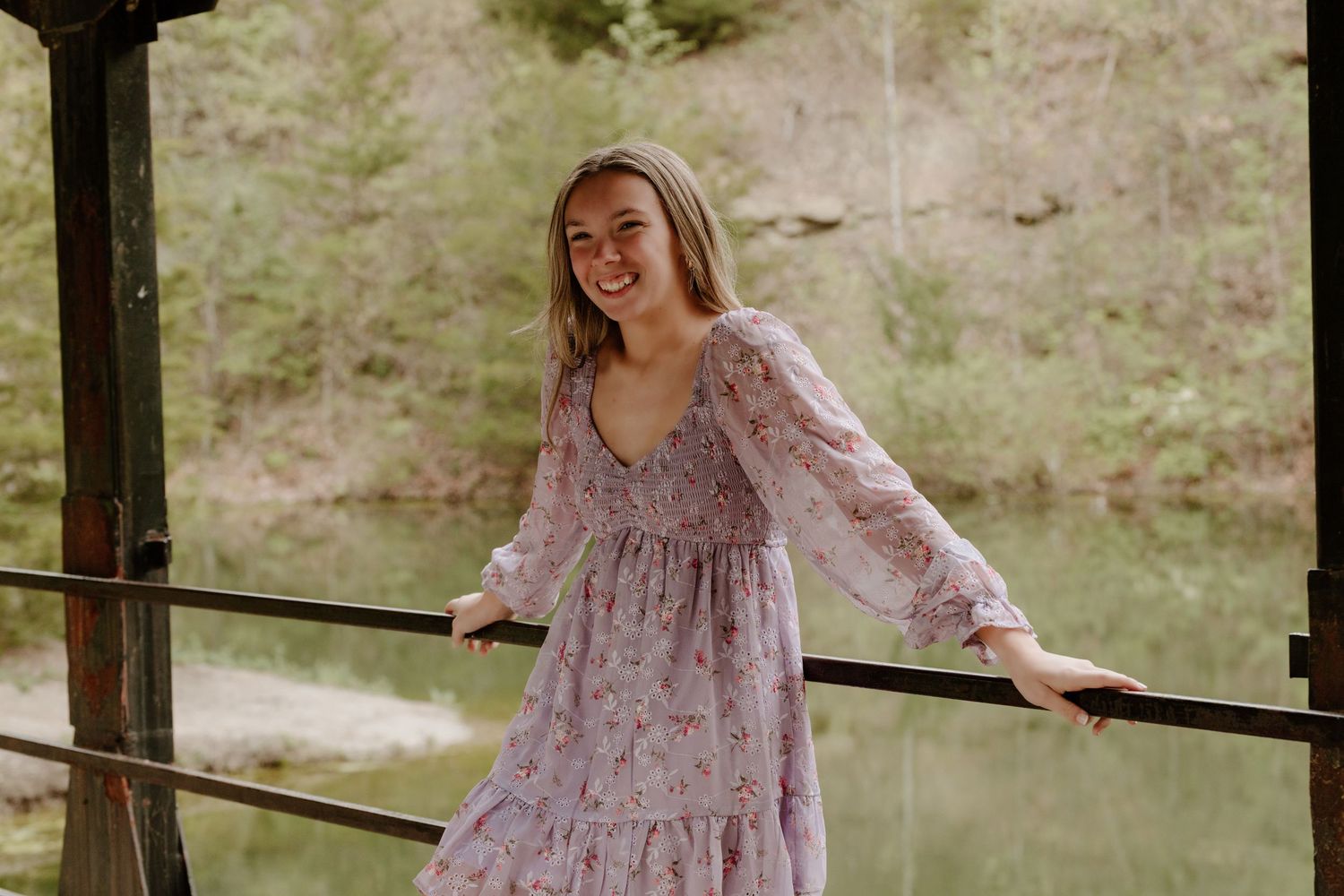 Someone wearing a floral mauve dress smiles and twirls on a lakeside deck during a sunny day.