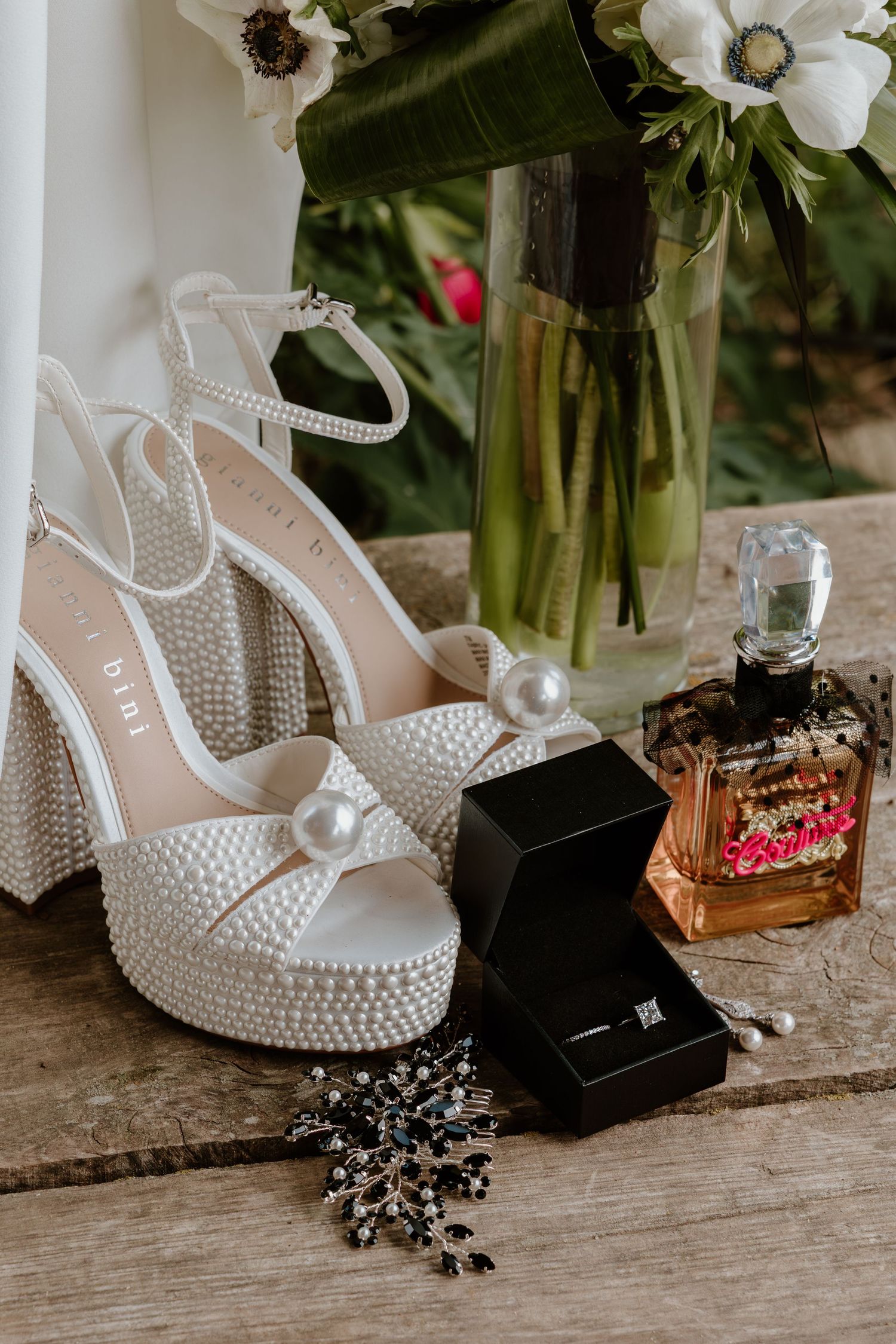 Pearl-embellished bridal heels displayed with a diamond ring box, floral brooch, and perfume on a wooden surface.