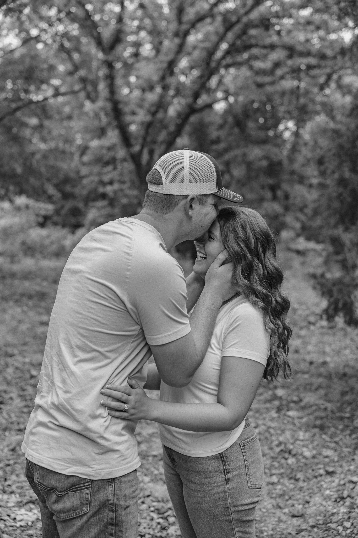 Black and white photo of a couple sharing a tender moment outdoors, man in cap gently holding womans face while she smiles.