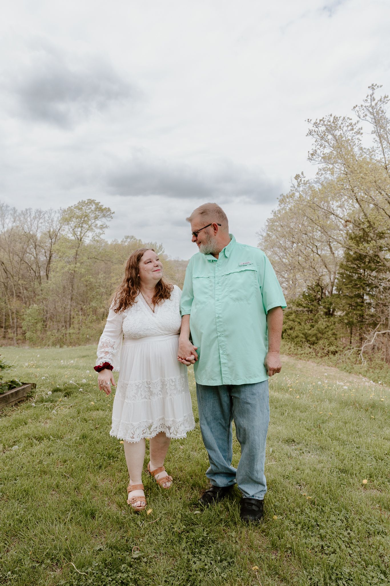 Couple holding hands and smiling at each other in a green meadow, woman in white dress, man in mint shirt.