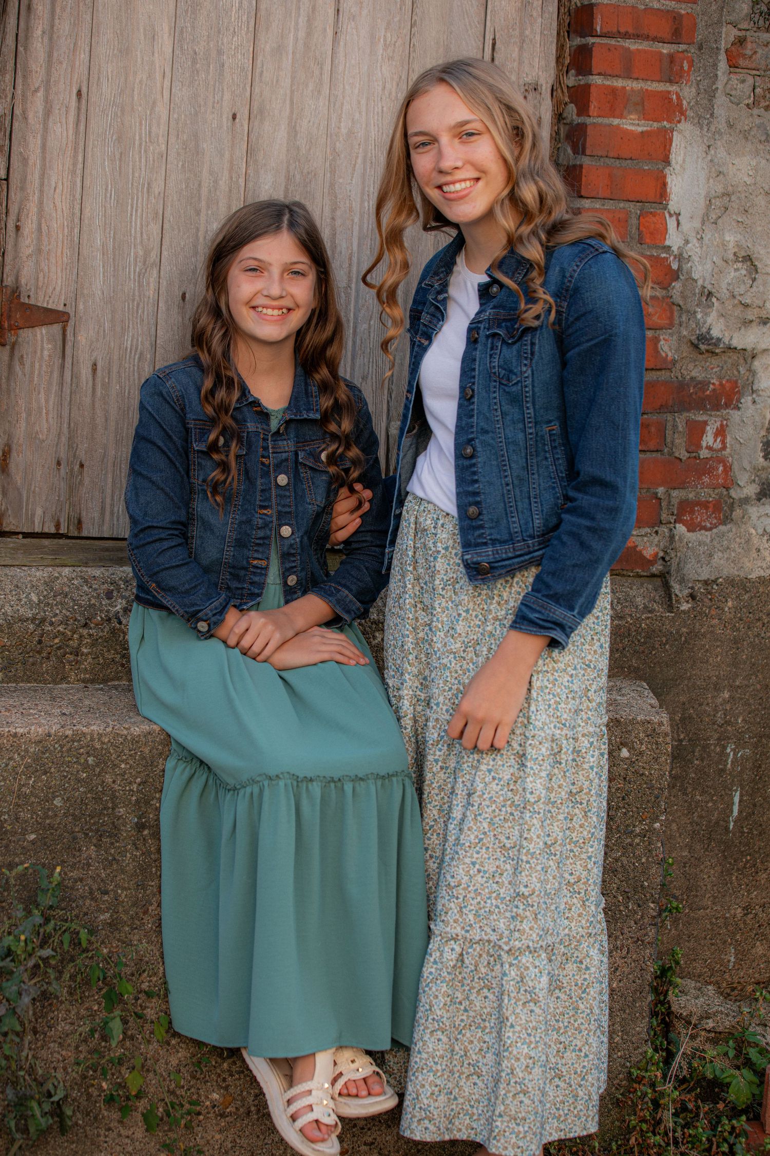 Two people in denim jackets and long skirts pose together against a weathered wooden barn wall.