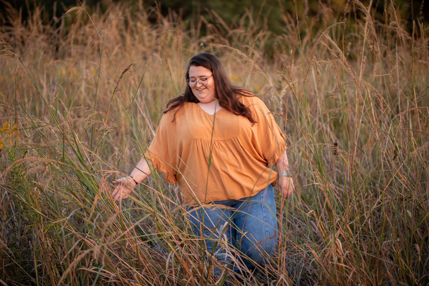 Person in orange sweater and jeans stands in tall grass during golden hour.