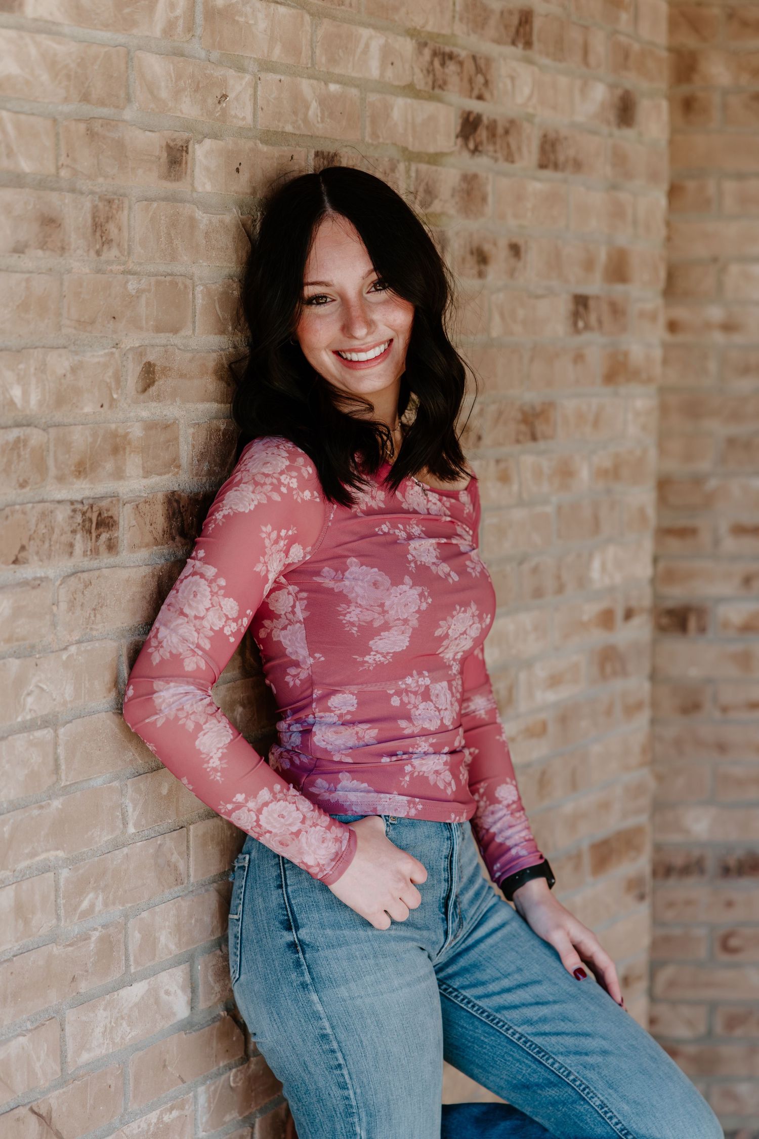A person in a pink floral mesh top and blue jeans poses against a brick wall with a warm smile.