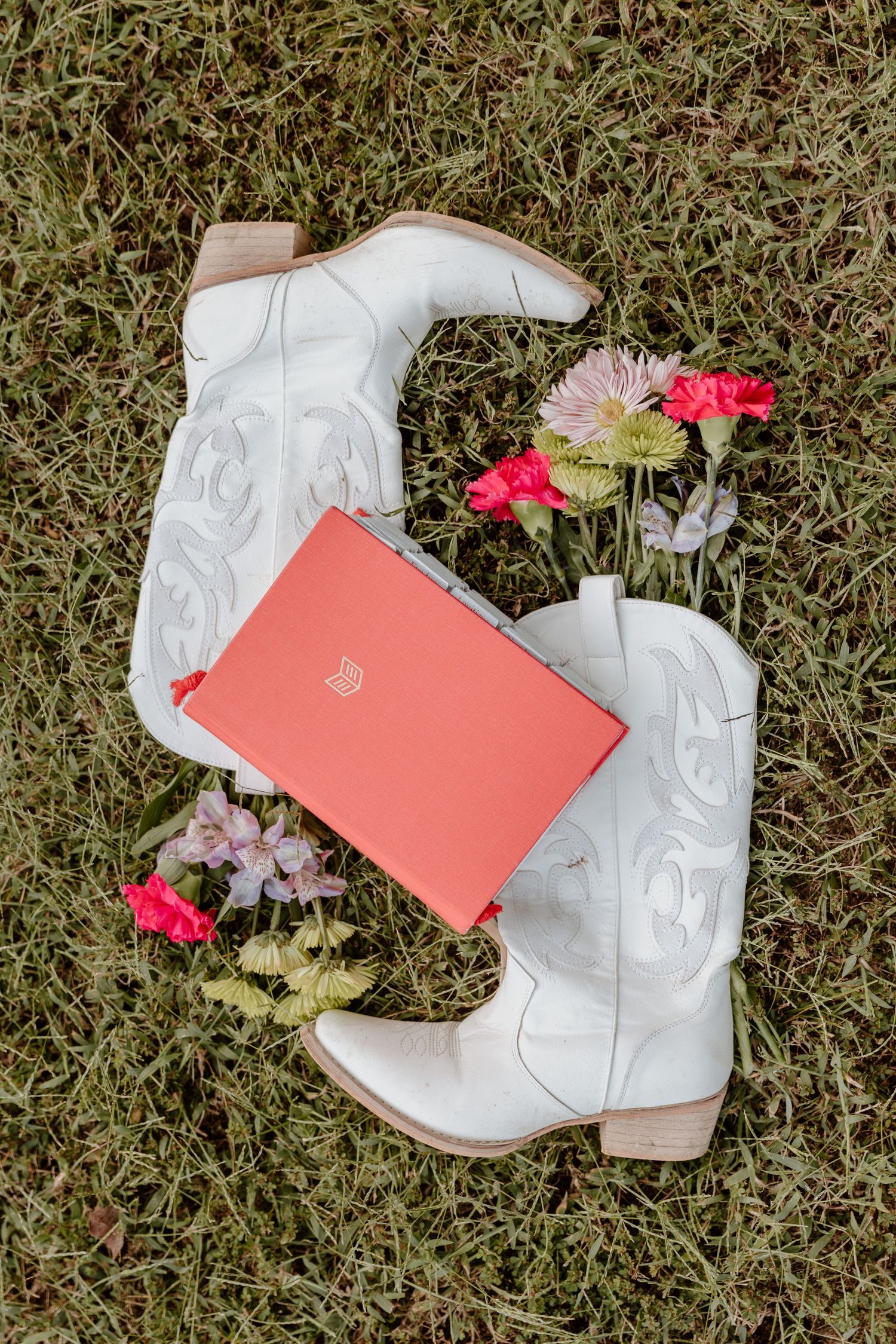 White leather cowboy boots with pink flowers and coral invitation cards arranged on grass in a wedding styled photoshoot.