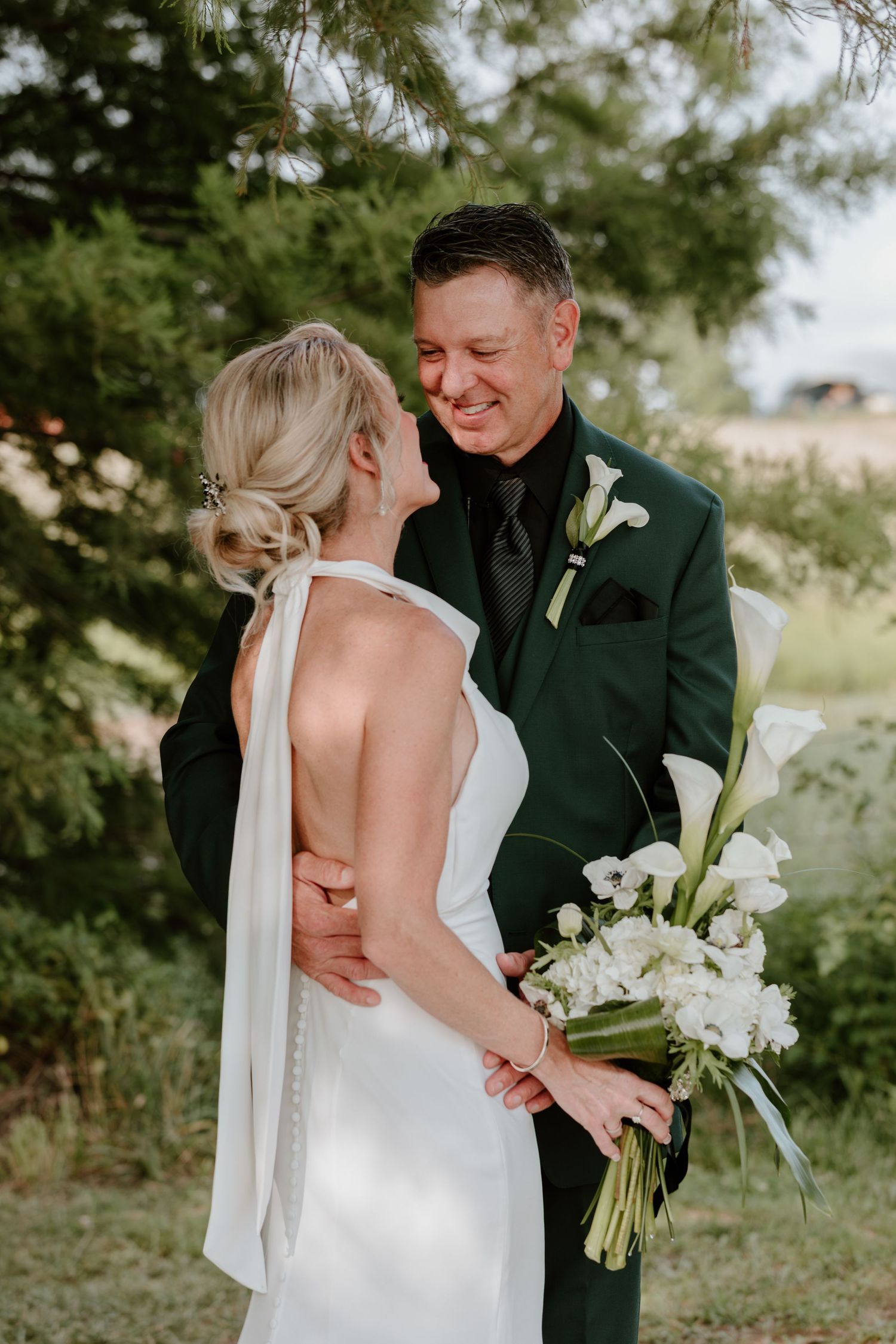 Smiling groom in dark green suit embraces bride in backless white gown while holding white calla lily bouquet outdoors.