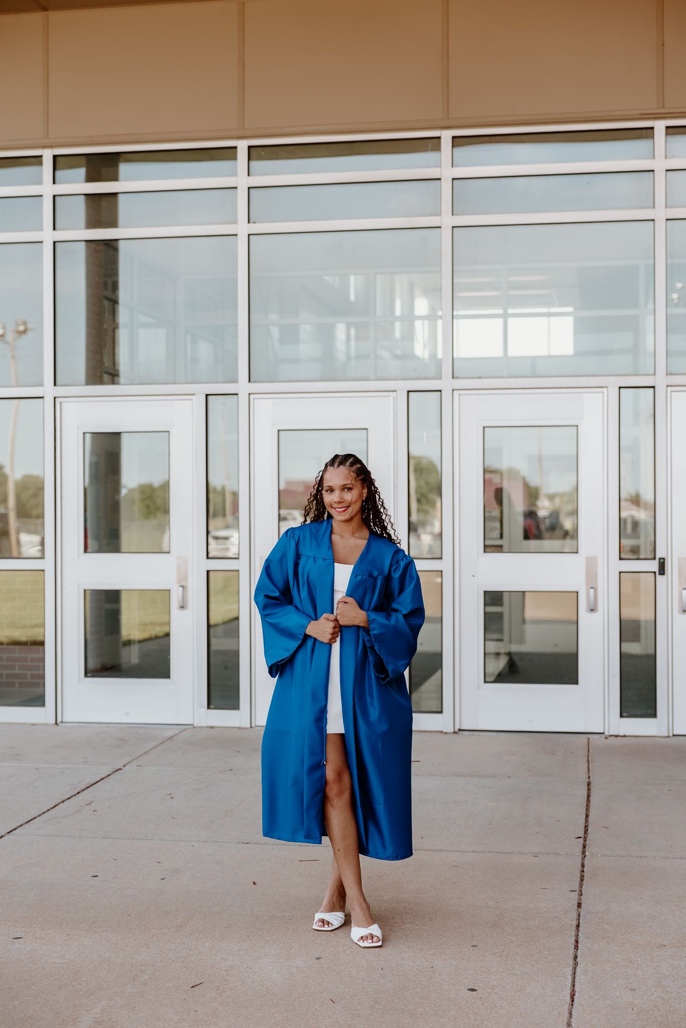 A smiling graduate in a blue graduation gown stands proudly outside school doors on a sunny day.
