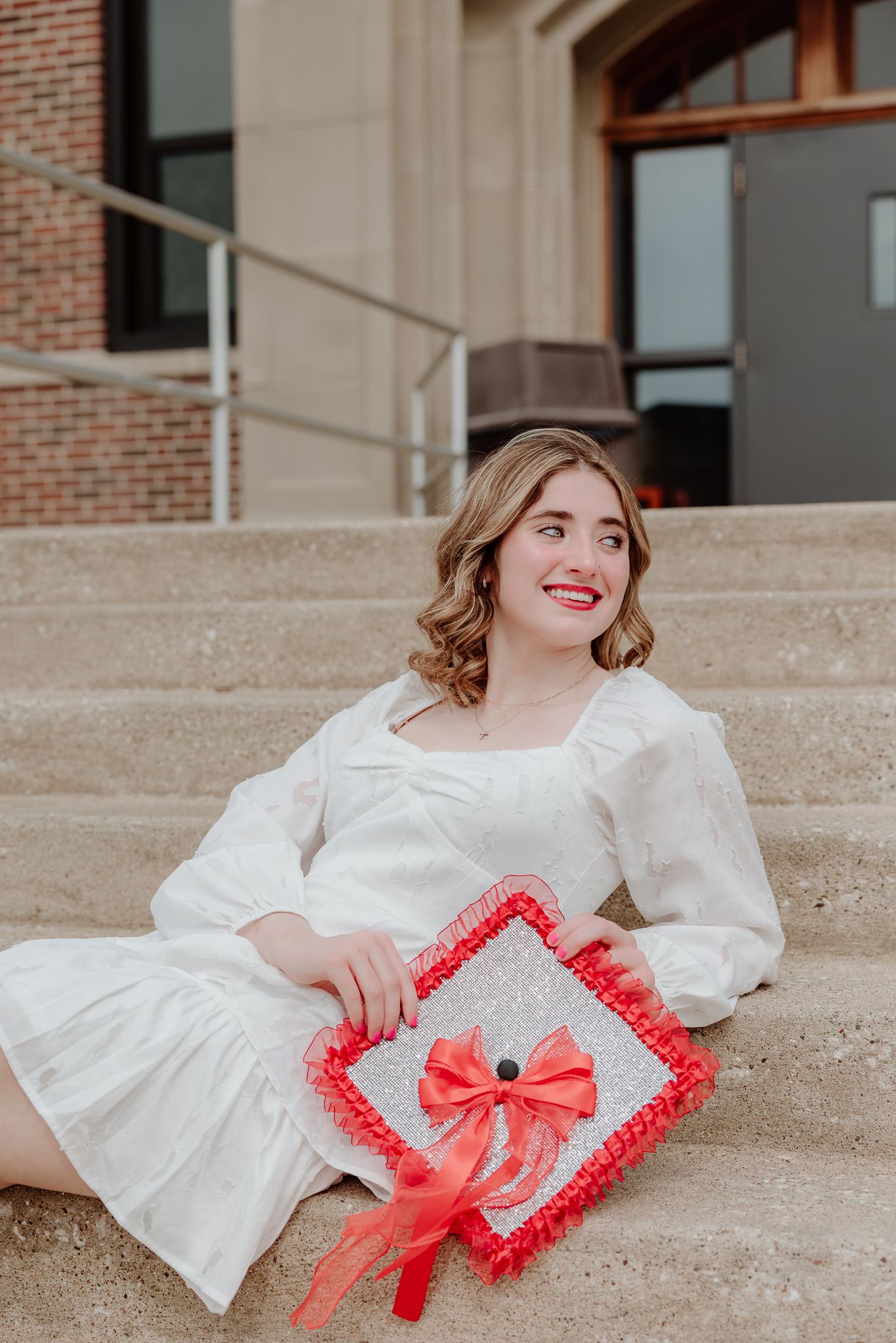 Young woman in white dress sitting on steps holding a decorated red graduation cap, smiling joyfully.