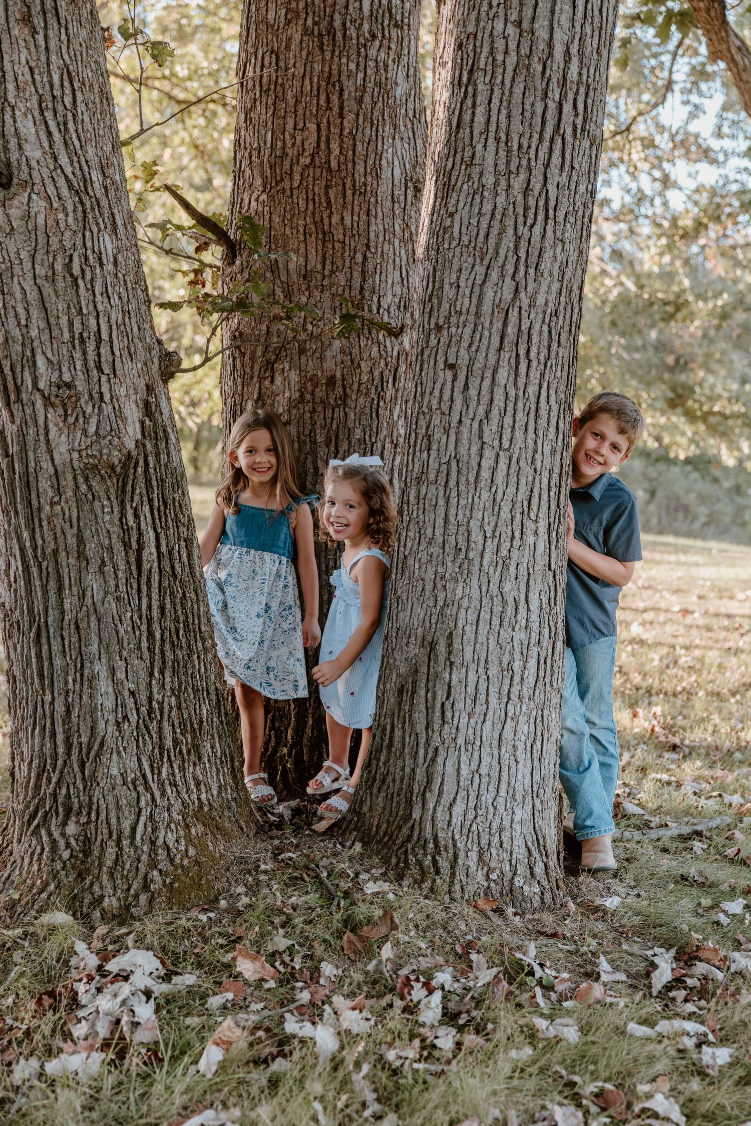Three children playfully pose around a large tree trunk in a sunlit autumn setting with fallen leaves on the ground.