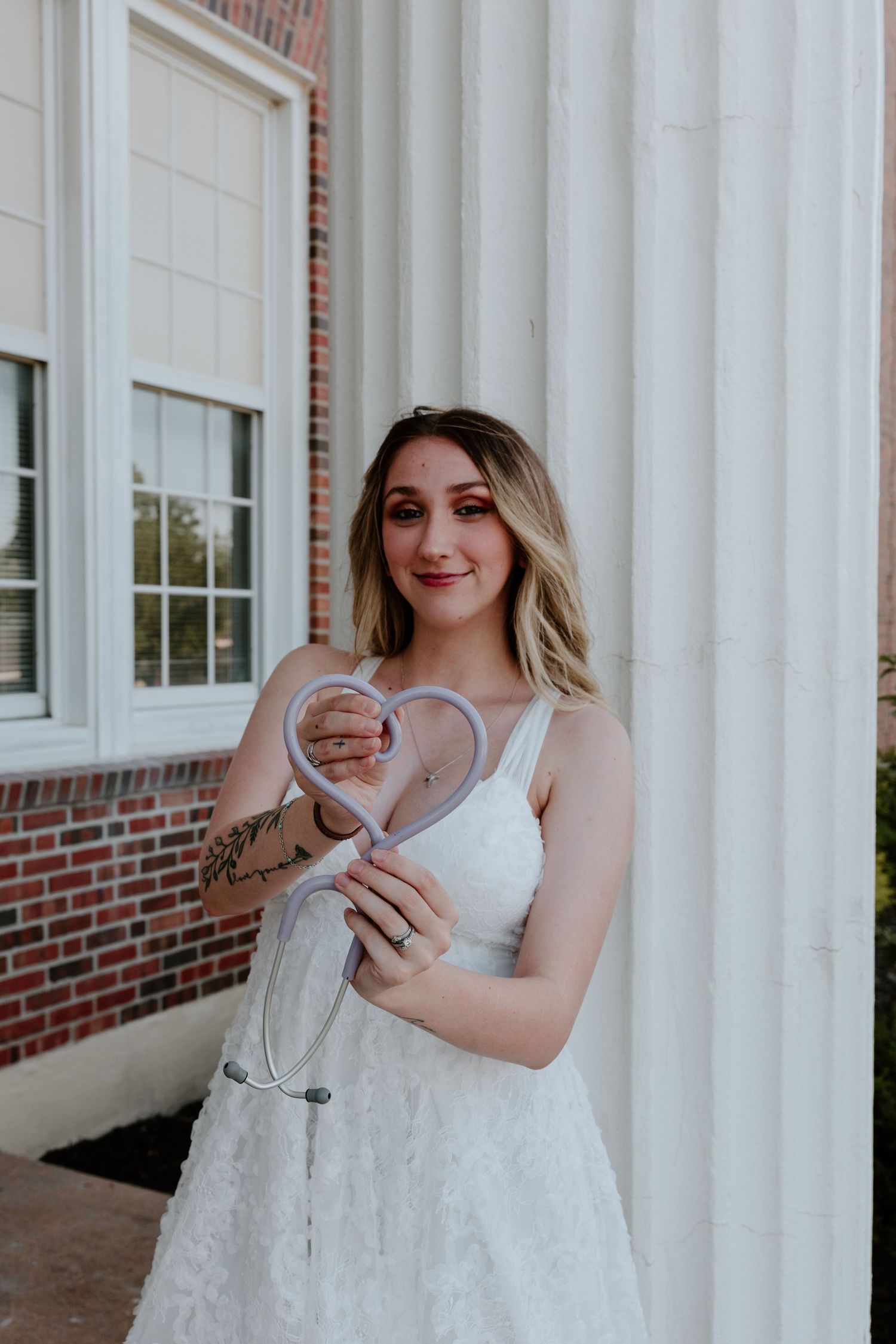A person in a white dress holds a heart-shaped sign while standing by white columns and brick building exterior.