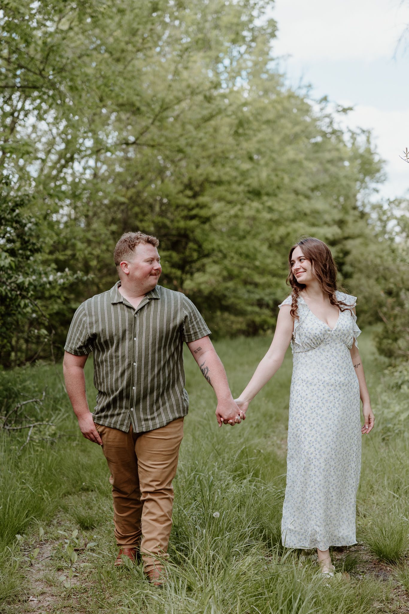 A couple holds hands and smiles at each other while walking through a lush green field during an outdoor engagement session.