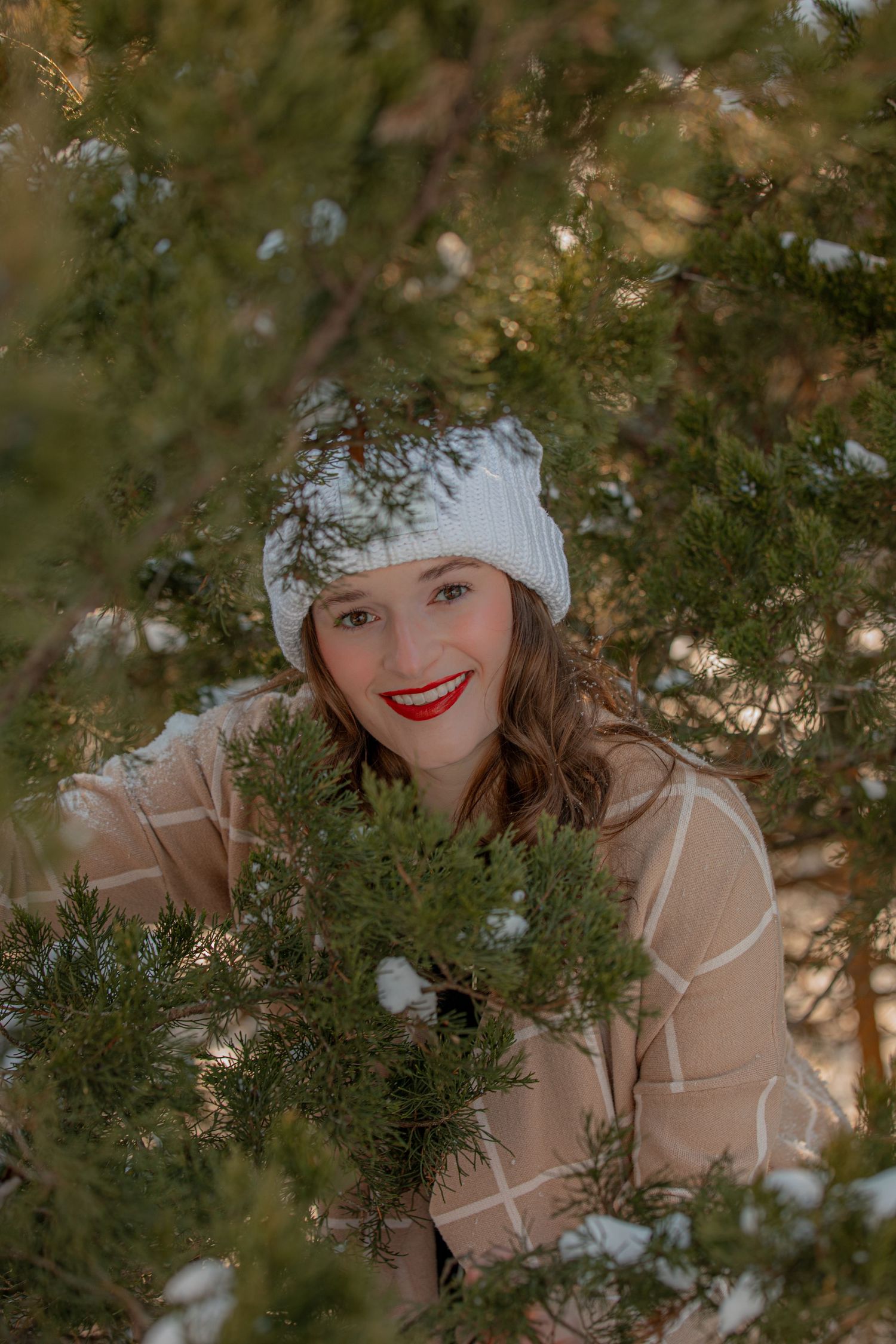 Winter portrait through evergreen branches with white knit hat and natural makeup.